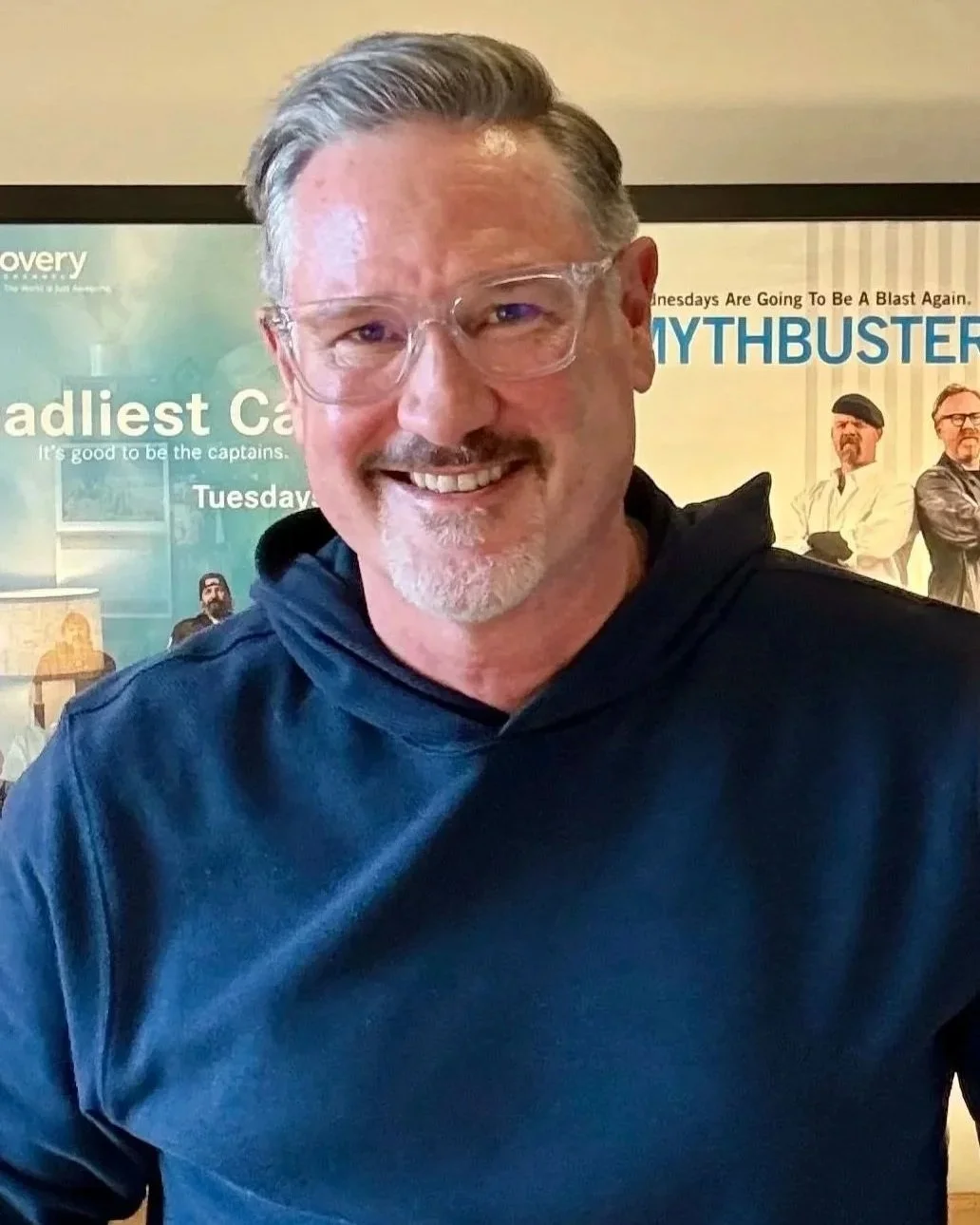 A man with glasses and gray hair smiling in front of a background with books and posters.