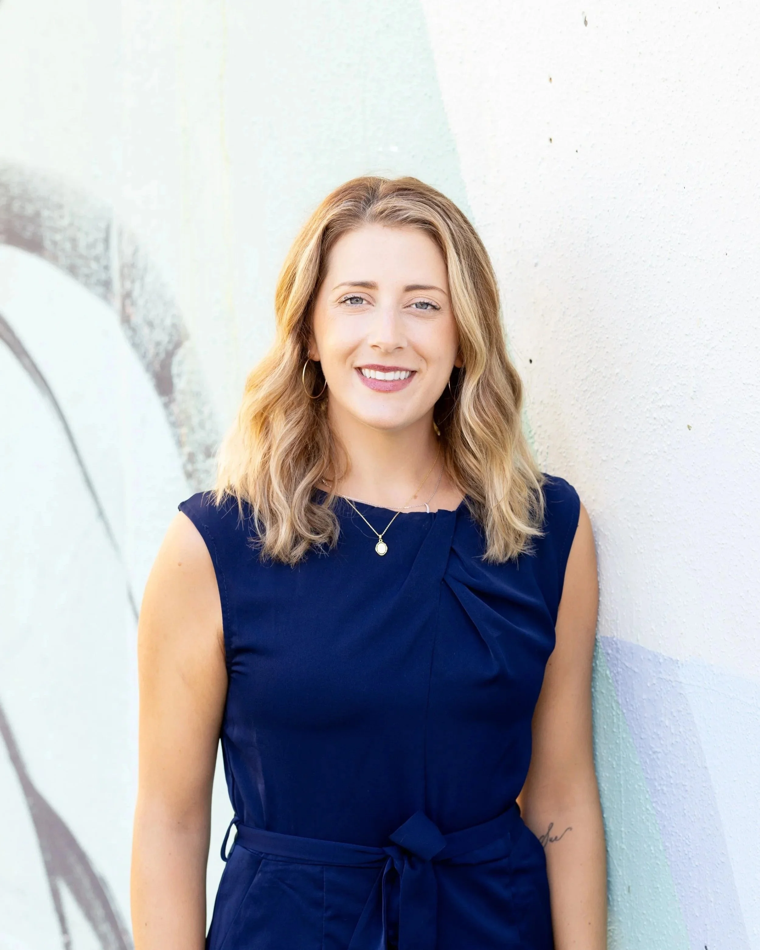 A young woman with blonde hair and blue eyes, smiling gently, wearing a white lacy top and small gold hoop earrings, standing outdoors in natural light.