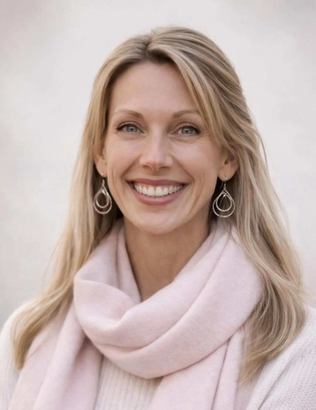 Woman with blonde, below the shoulder length hair wearing blue jeans, a white turtleneck shirt with a deep wine colored blazer standing outside under scenic trees