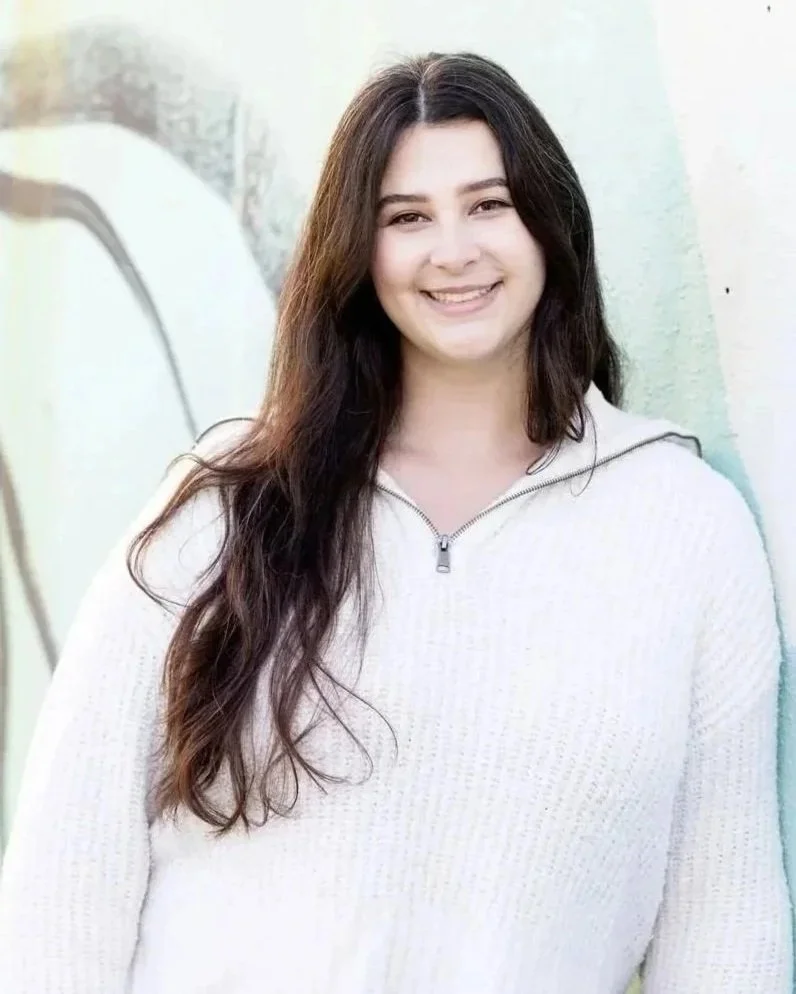 A young woman with long dark hair smiling, wearing a cream-colored zip-up sweater, standing against a light-colored wall with some abstract art in the background.