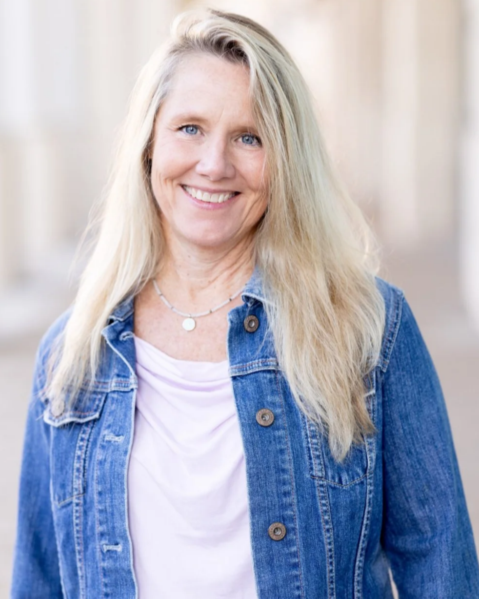 A woman with long blonde hair smiling outdoors, wearing a pink shirt and layered necklaces.