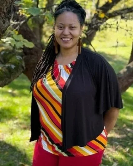 Woman smiling outdoors with trees in the background, wearing a colorful striped top, black jacket, and red pants.