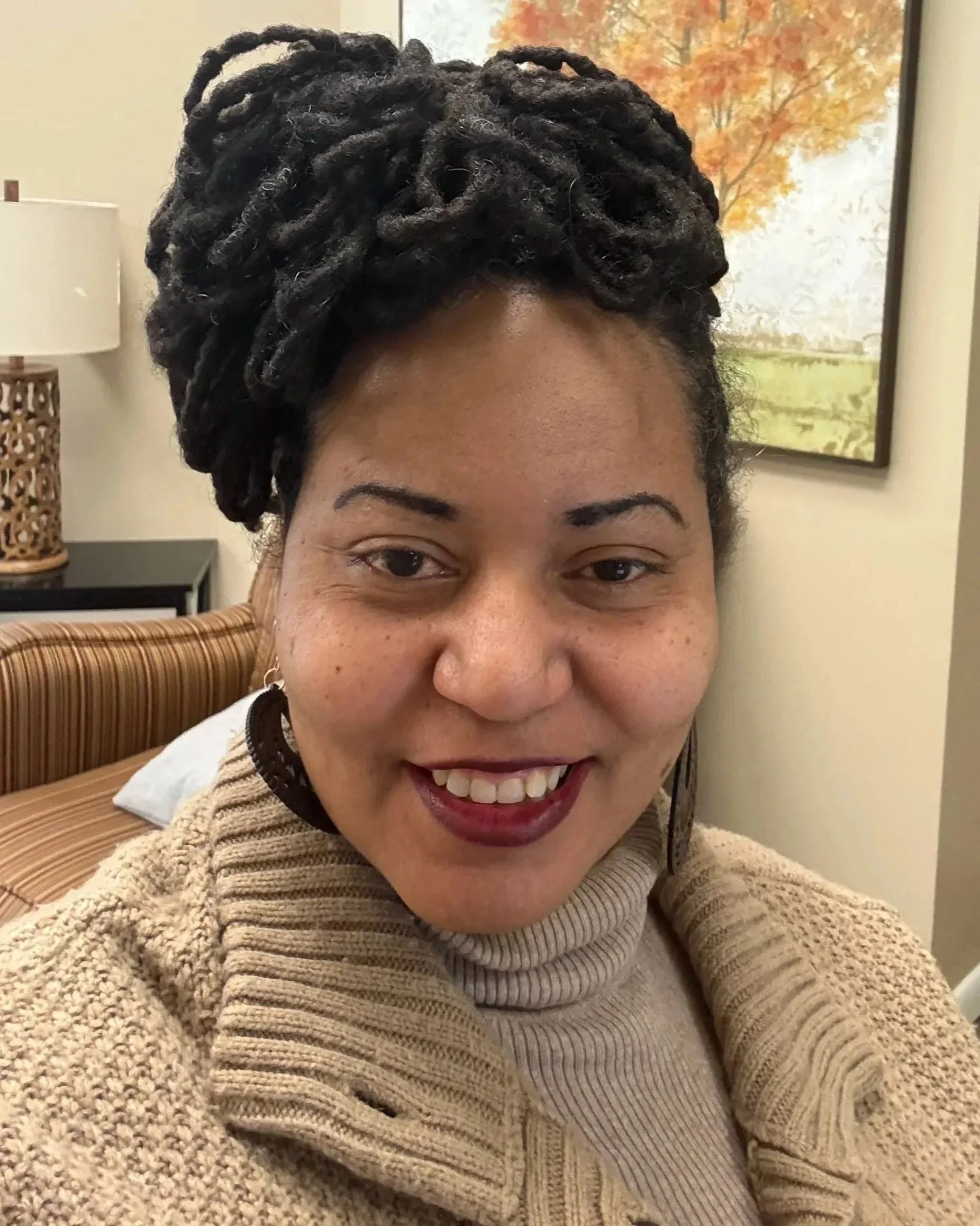 Woman smiling outdoors with trees in the background, wearing a colorful striped top, black jacket, and red pants.