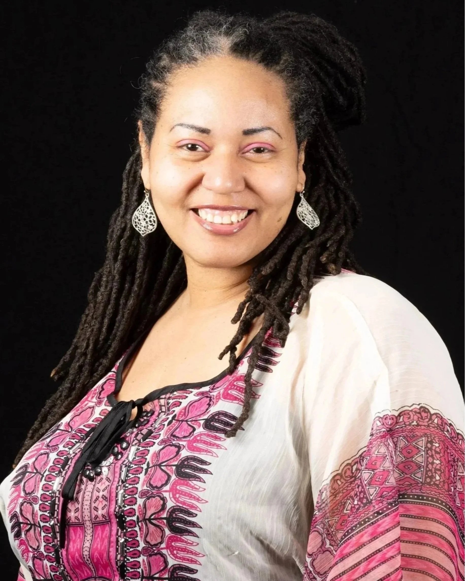 A woman standing outdoors near a tree in a park during daytime, smiling, wearing a colorful striped top, a black shawl, red pants, and large earrings.