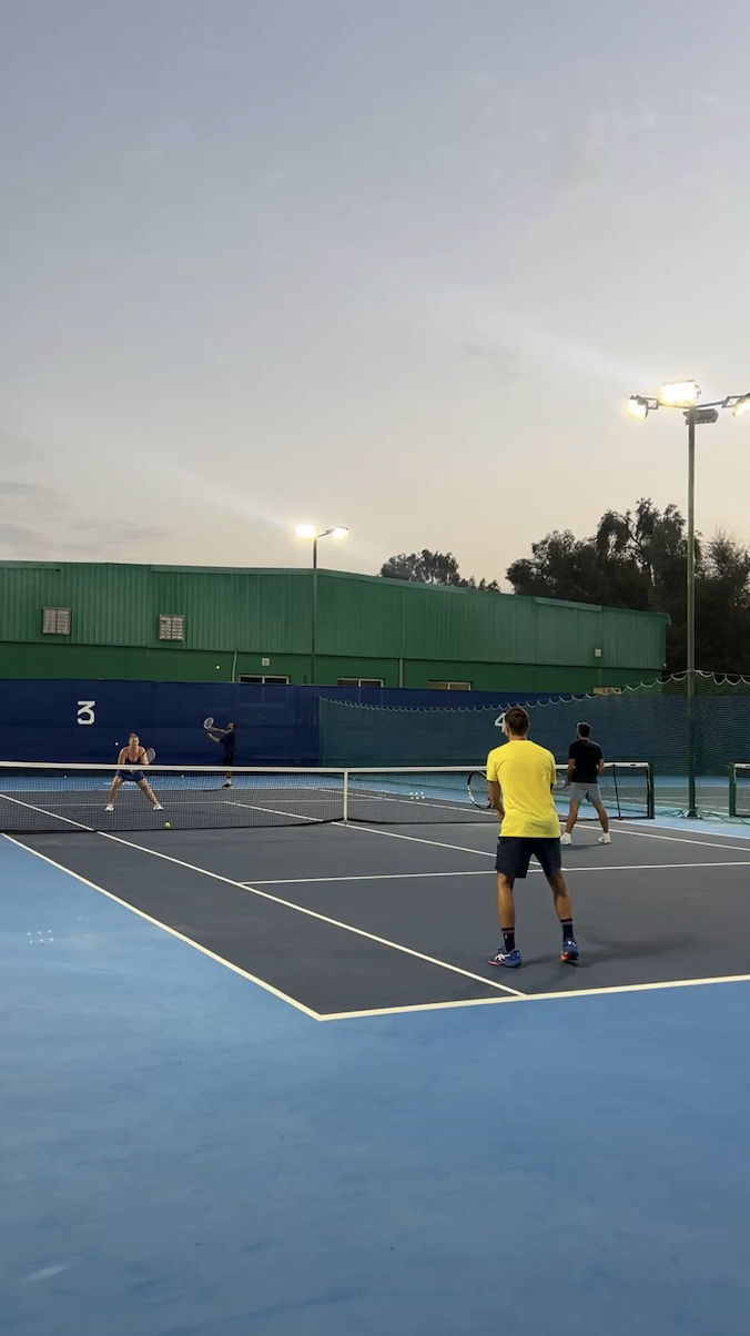 Four young people playing tennis on an outdoor court during evening, with green industrial building and tall lights in the background.