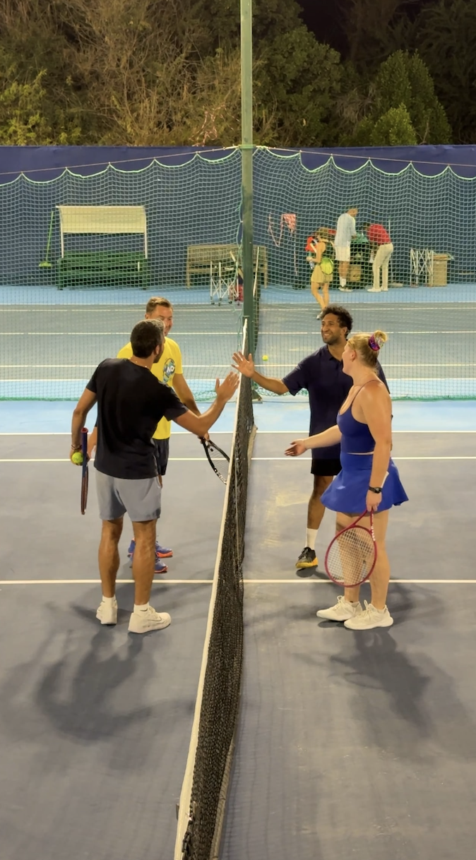 Four tennis players, two men and two women, standing on a tennis court at night, shaking hands over the net after a match. The court is surrounded by a fence and trees, with some people seen in the background near the back fence.