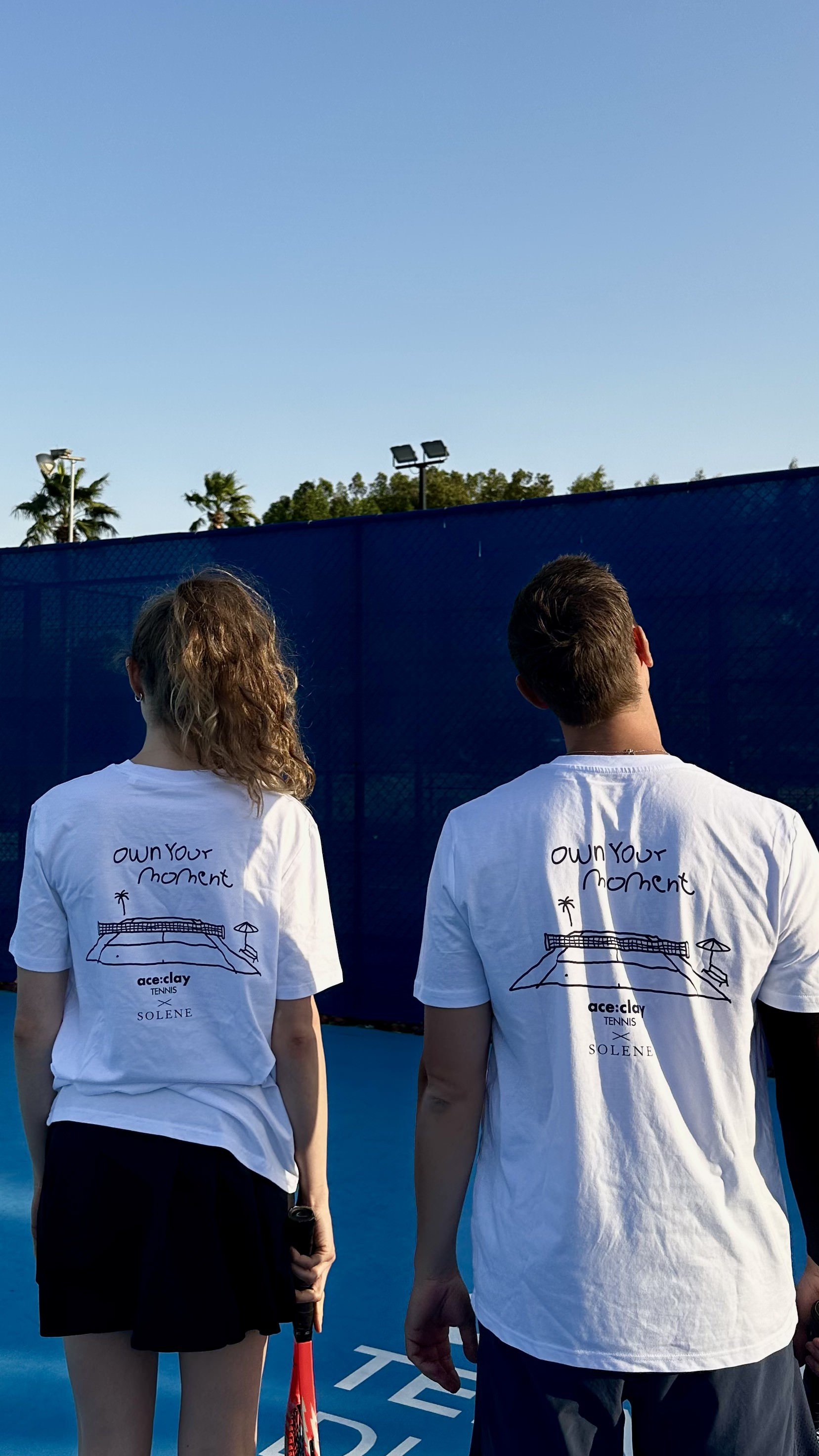 Two people seen from behind, wearing matching white t-shirts with a drawing of a tennis court and the words 'own your moment,' standing on a blue tennis court near a dark blue fence under a clear sky.