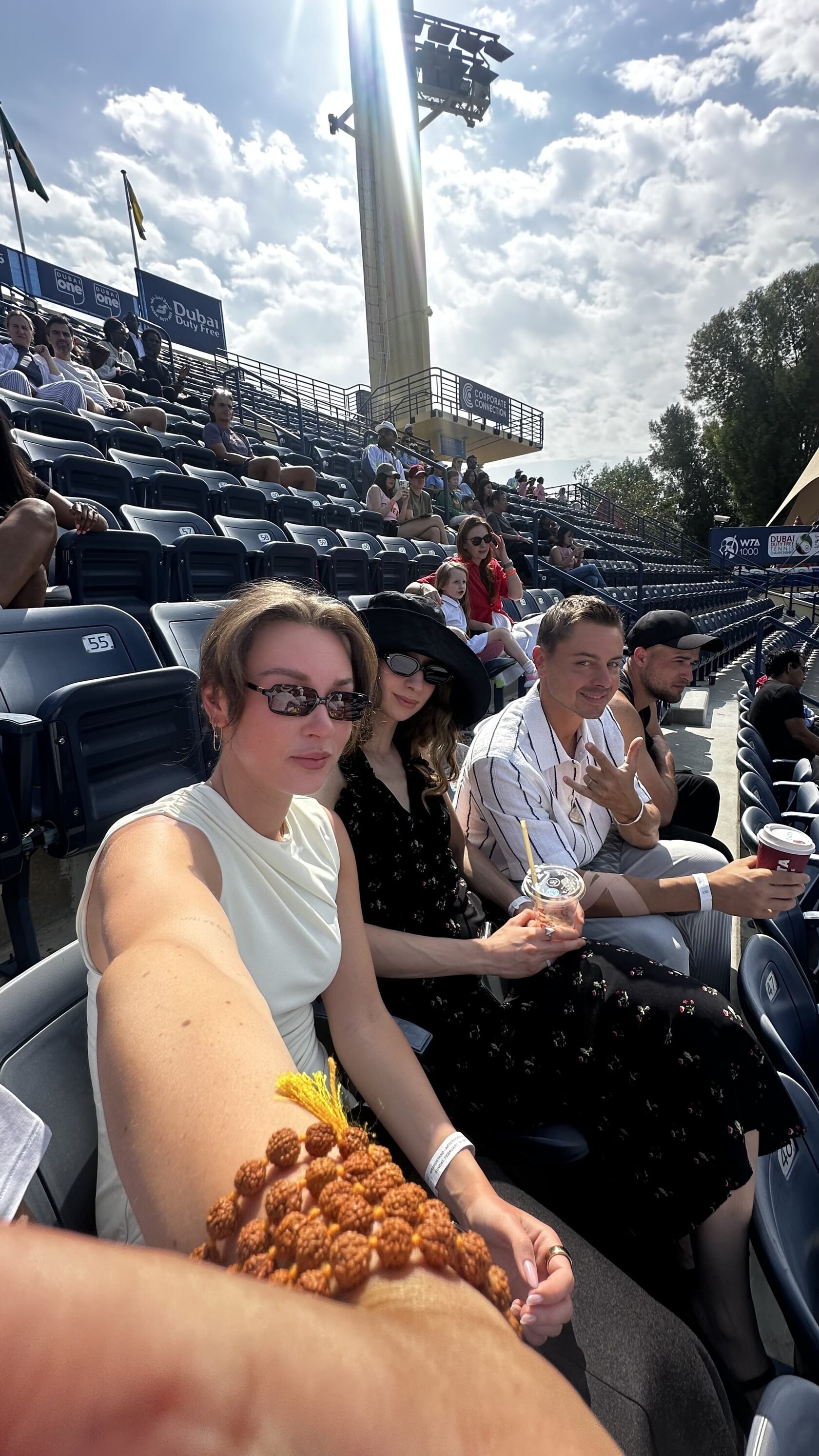 People sitting in stadium seats at an outdoor tennis event, with a few holding drinks and wearing casual and stylish clothing, under a partly cloudy sky.