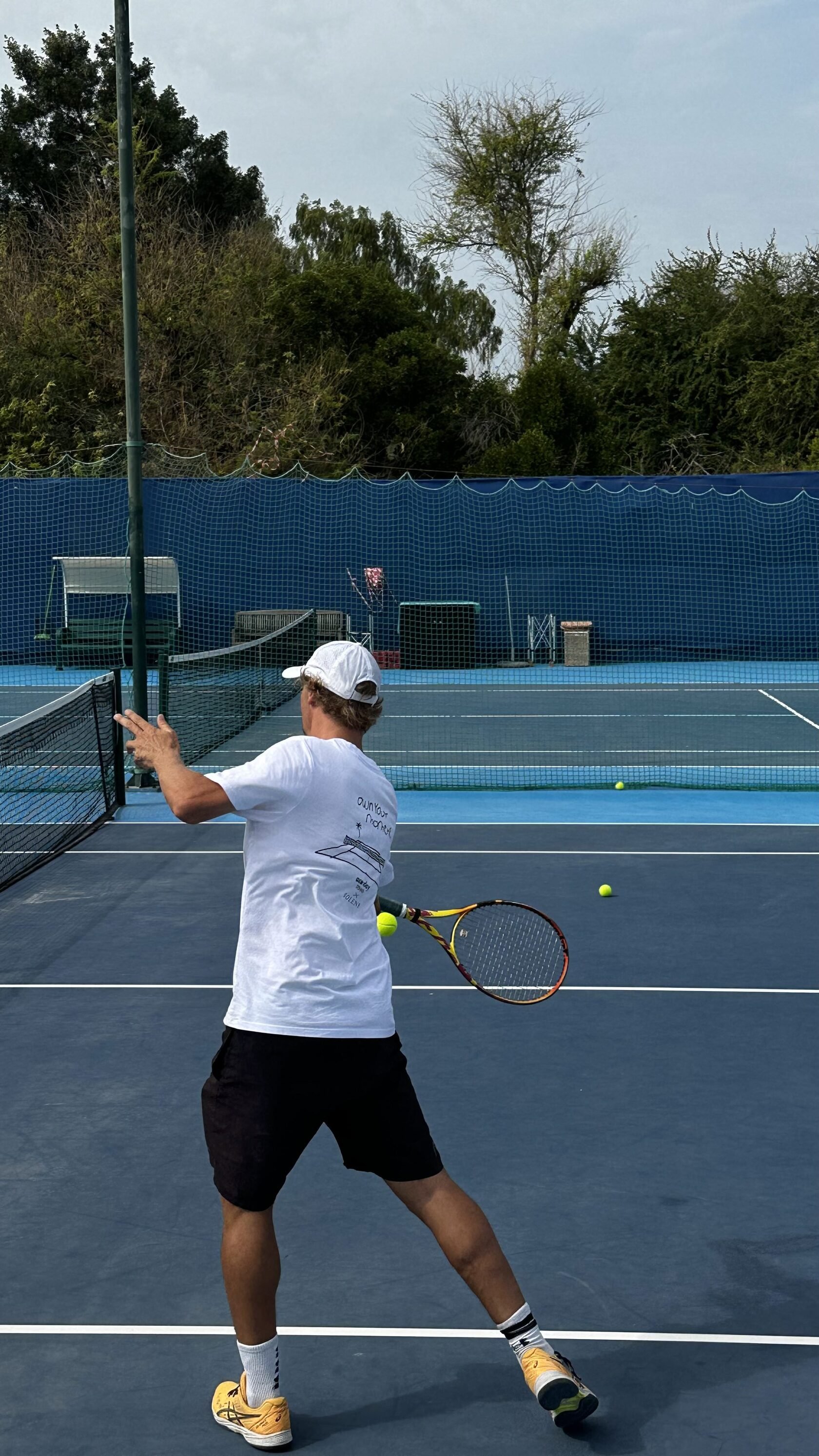 A man playing tennis on an outdoor court with a blue surface, holding a tennis racket, preparing to hit a tennis ball. The court is surrounded by a dark blue fence, and there is a net in front of him.