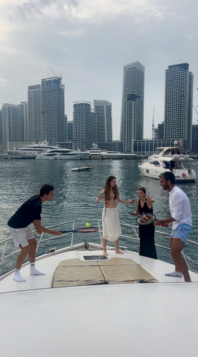 Four people playing tennis on the deck of a yacht with a city skyline and yachts in the background.