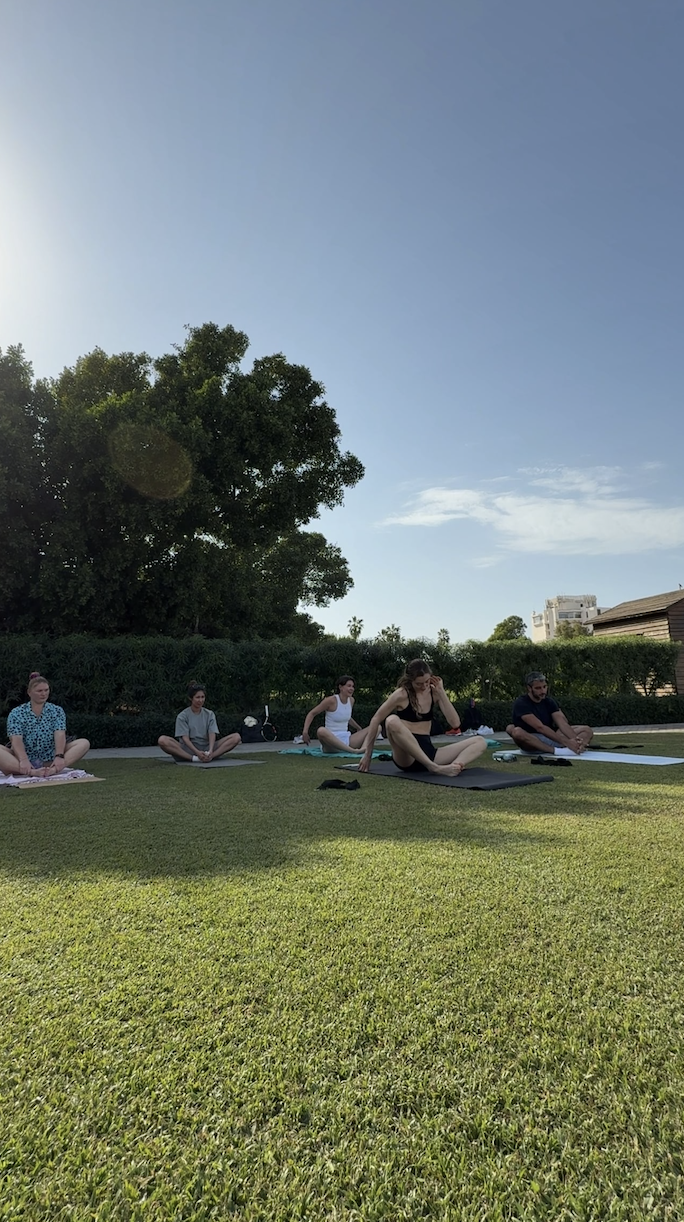 A group of people participating in an outdoor yoga class on a sunny day, sitting on yoga mats on a grassy area with trees and buildings in the background.