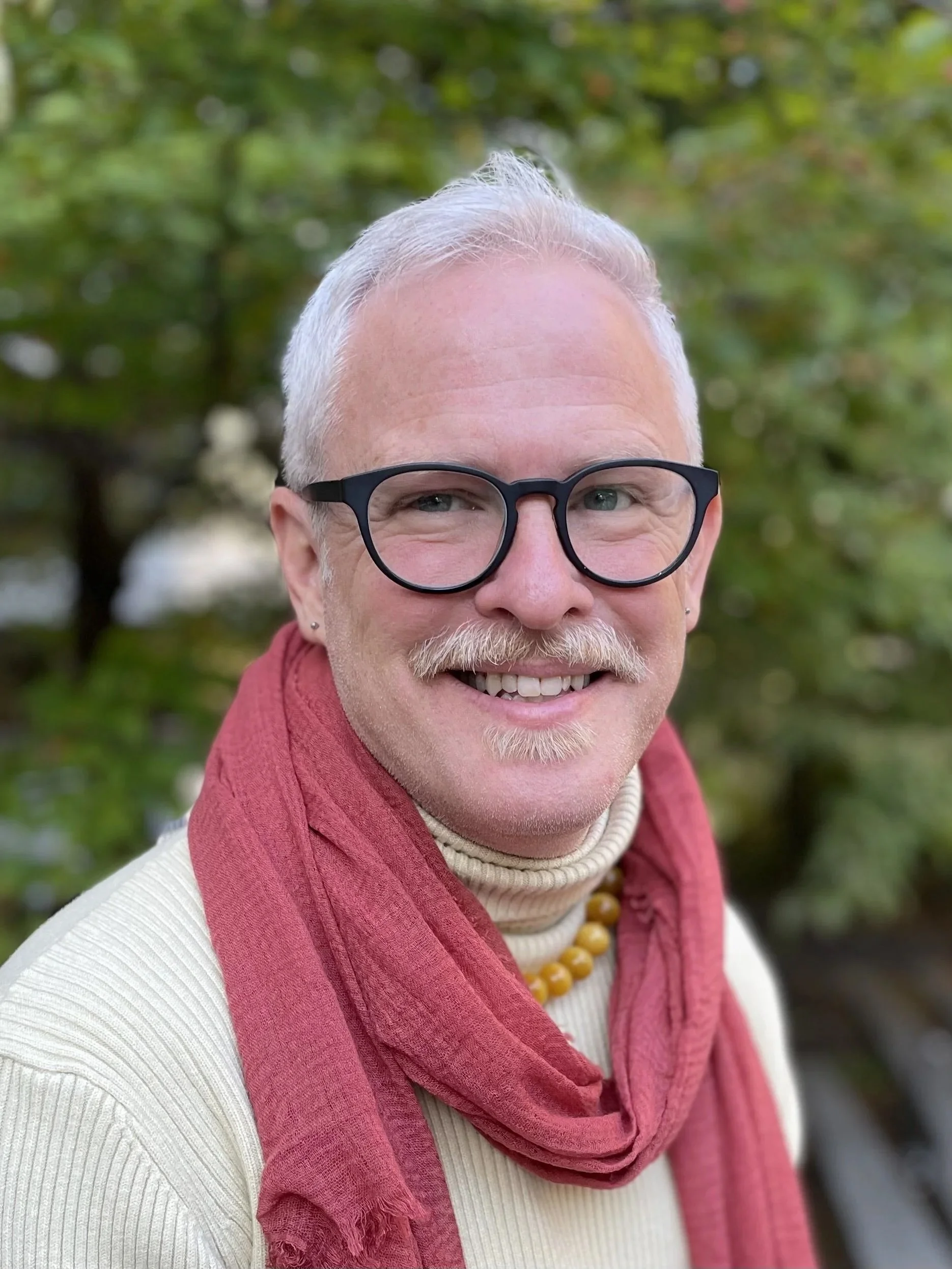 A headshot of Monroe wearing a cream-colored turtleneck and a red scarf outdoors with blurred green foliage in the background.