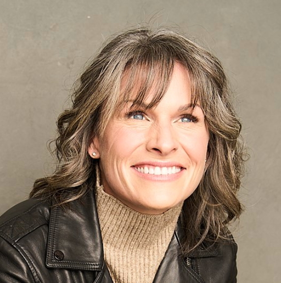 Smiling woman with wavy brown hair, blue eyes, wearing a beige turtleneck sweater and black leather jacket, posing against a neutral background.