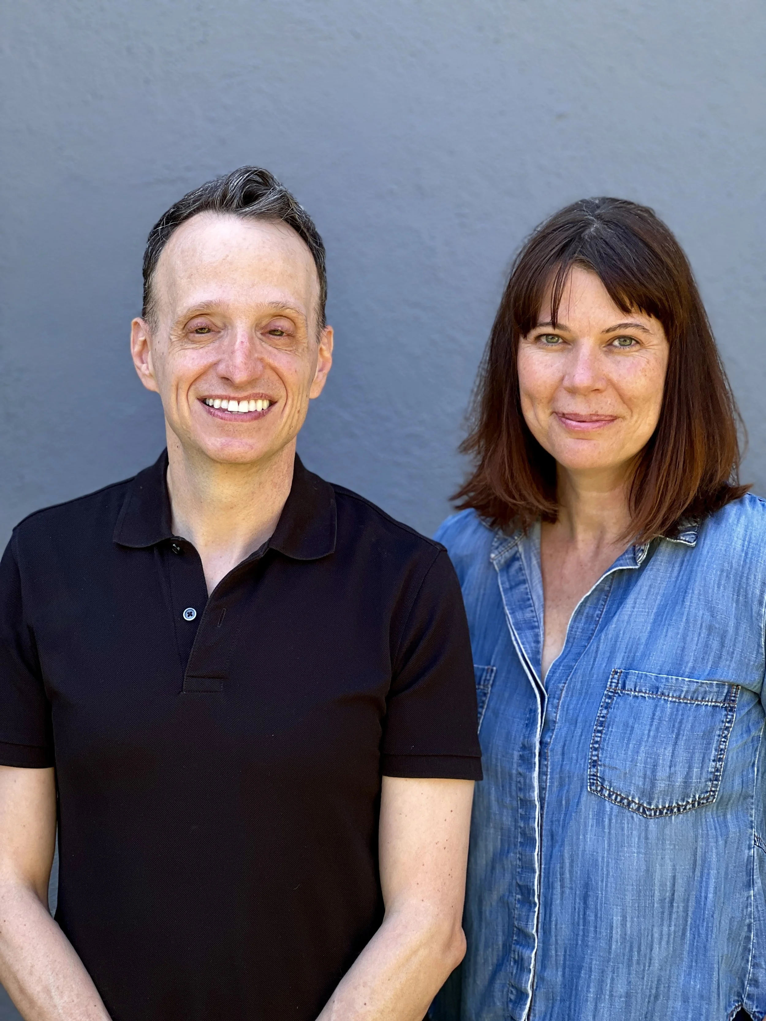 A man and woman standing side by side against a plain gray wall, both smiling at the camera.