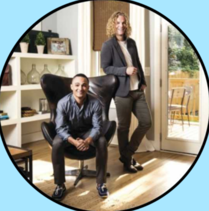 A woman and a man in a modern living room, with the woman standing and the man sitting in a black ergonomic chair, smiling at the camera. The room has white shelves with decorative items, a glass door leading outside, and natural light shining in.