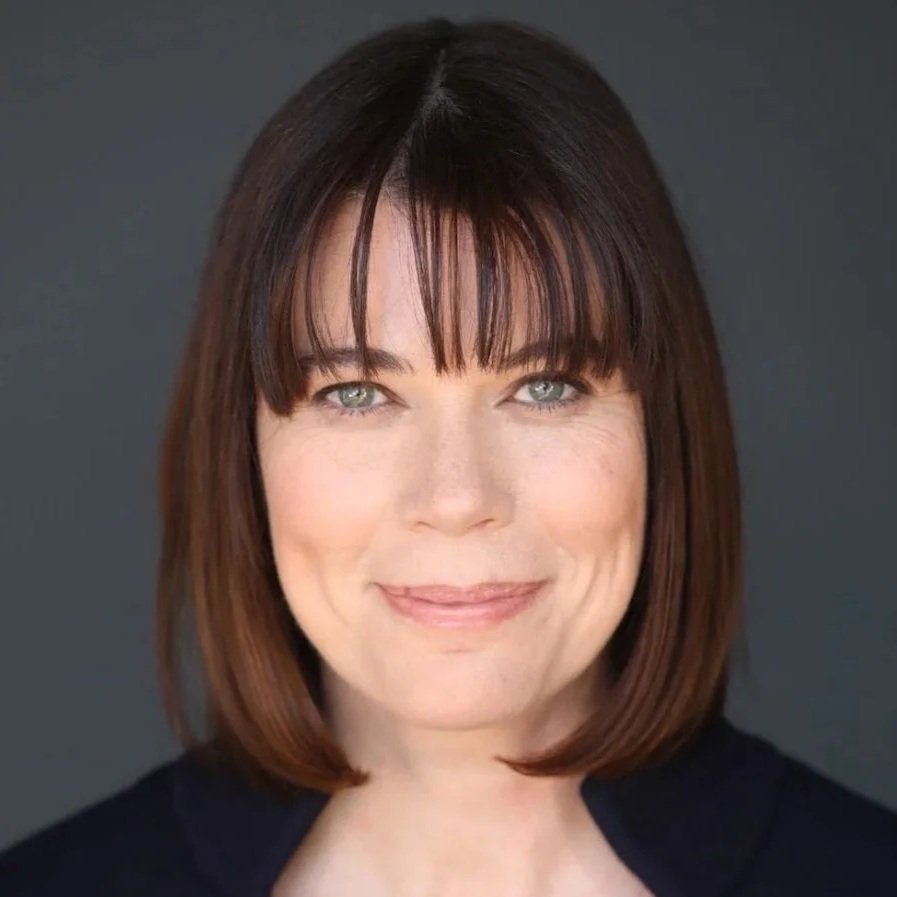 A woman with shoulder-length brown hair and bangs, smiling with light makeup, wearing a dark top, against a dark background.