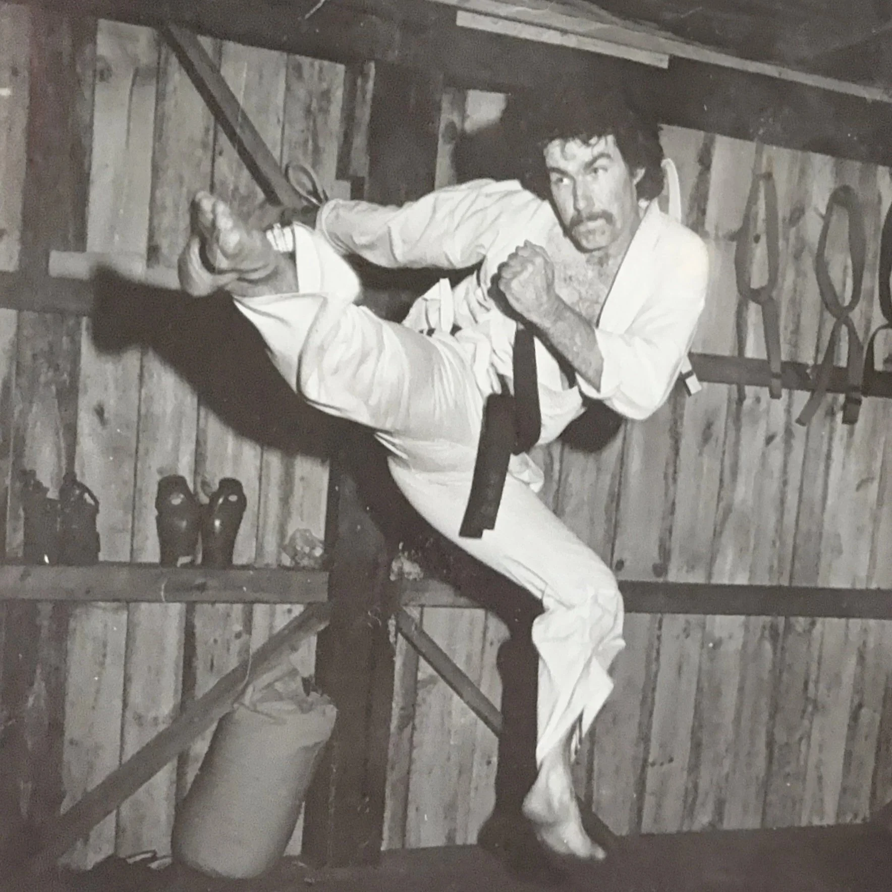 A man practicing martial arts in a dojo, wearing a traditional gi and black belt, in mid-air executing a high kick.