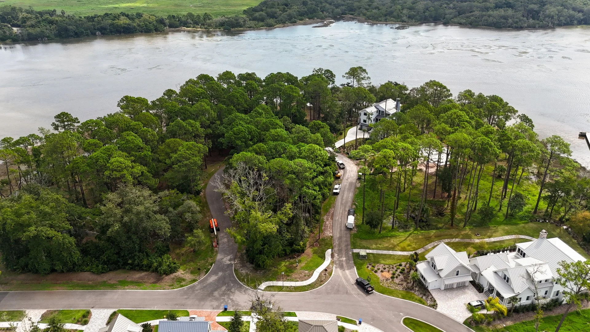 Aerial view of a residential neighborhood near a large body of water, with lush green trees, winding roads, and several houses, including one prominent white house with a metal roof.