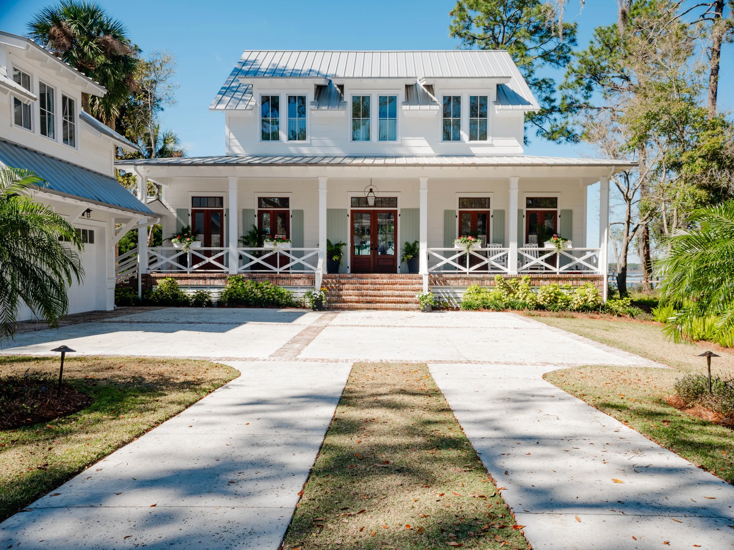 Crane Island Home. A white two-story house with a metal roof and a front porch, surrounded by a garden with green shrubs and trees, under a clear blue sky.