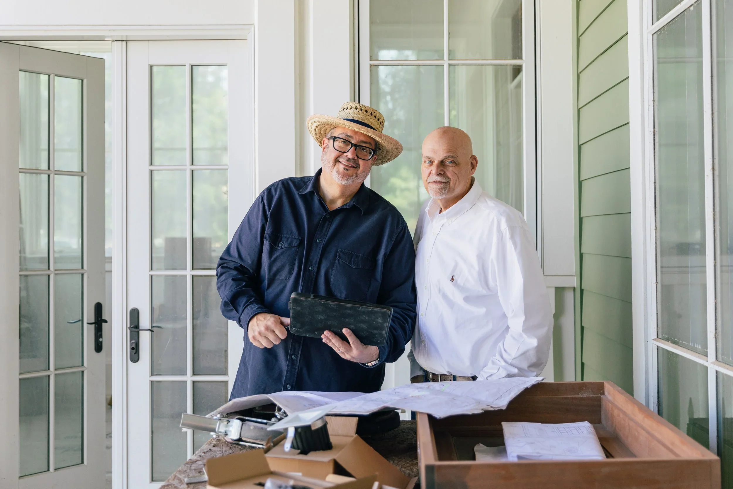 Gordon & Johnson Interior Design - Crane Island. Two men standing on a porch, smiling, with construction plans and tools on a table in front of them.