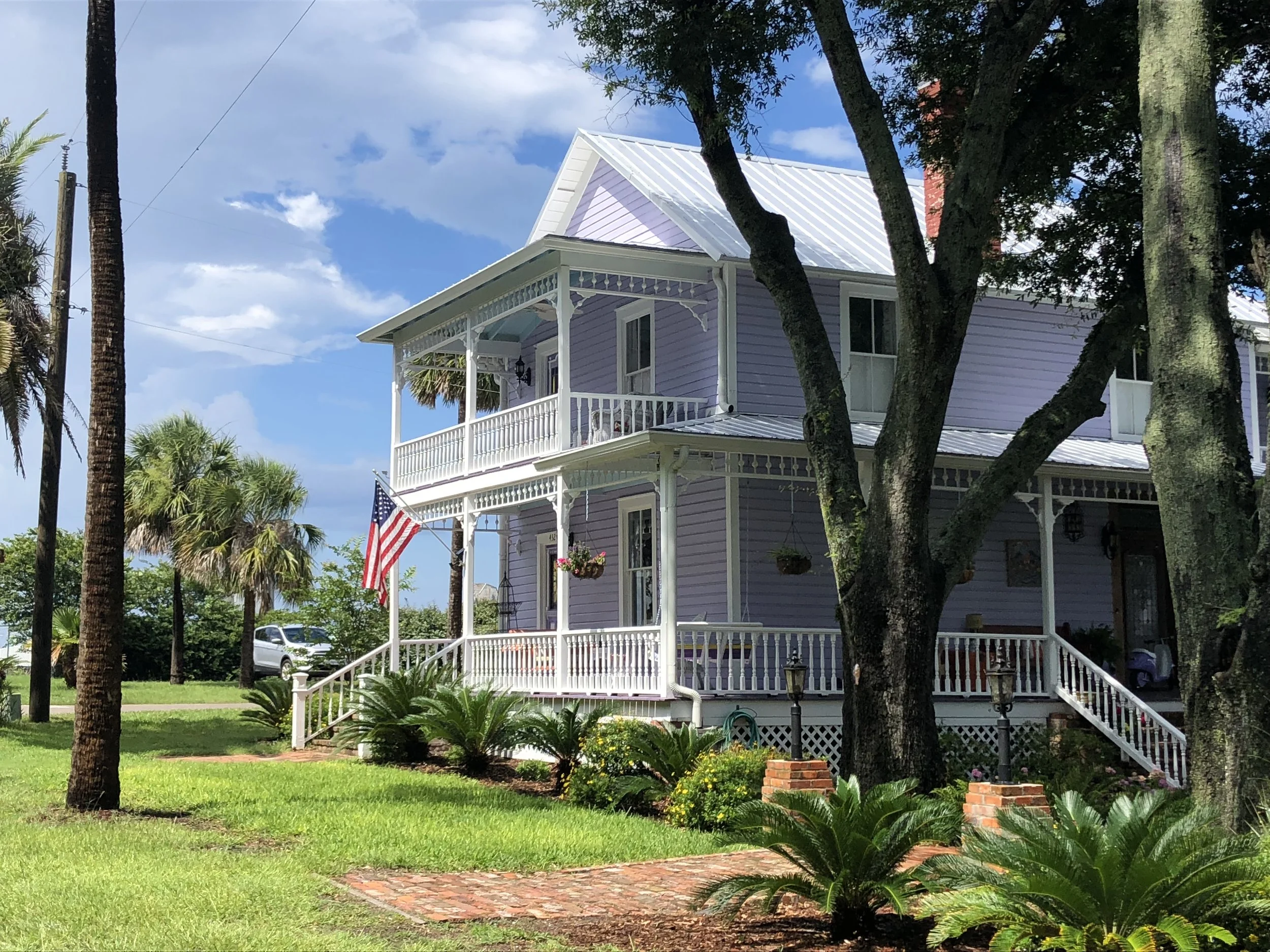 A two-story purple house with white trim and a metal roof, surrounded by lush greenery and trees, with a porch and steps leading up to the front door, and an American flag hanging from a pole on the porch. Downtown Fernandina Beach, FL - Examples of