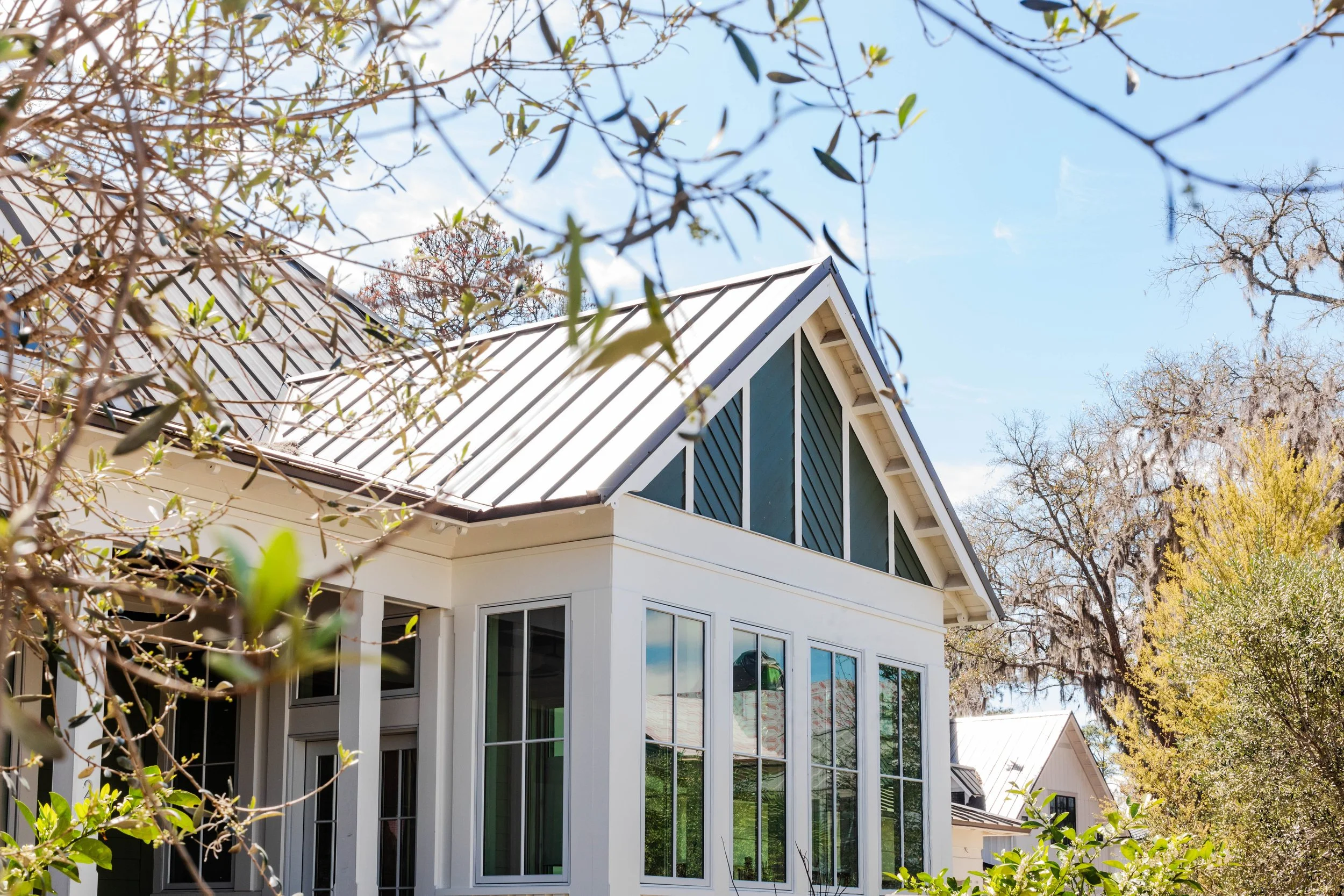 Historical Concepts - Crane Island. View of a modern house with large glass windows and a metal roof, surrounded by trees with some leaves and branches in the foreground, under a clear blue sky.
