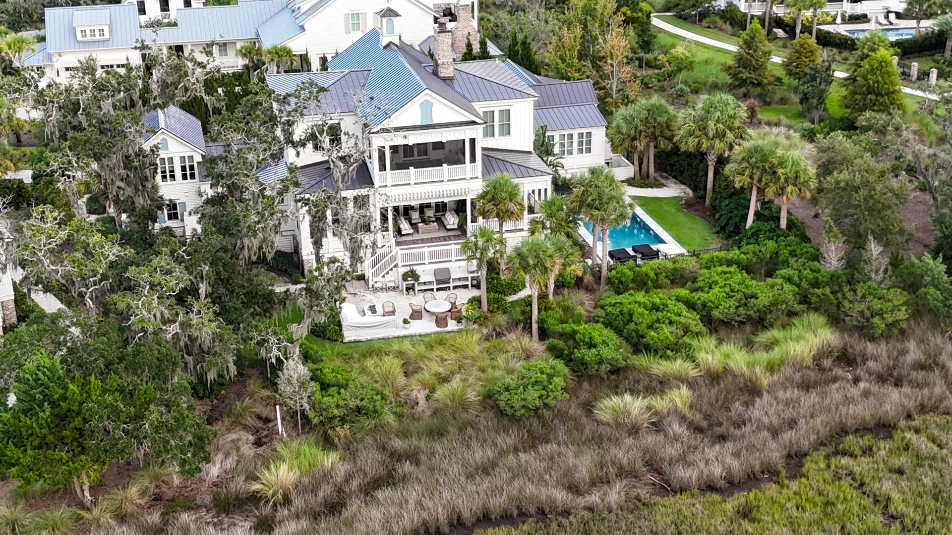 An aerial view of a large white house with a blue metal roof, surrounded by lush greenery, trees, and a backyard with an outdoor seating area, a swimming pool, and landscaping.