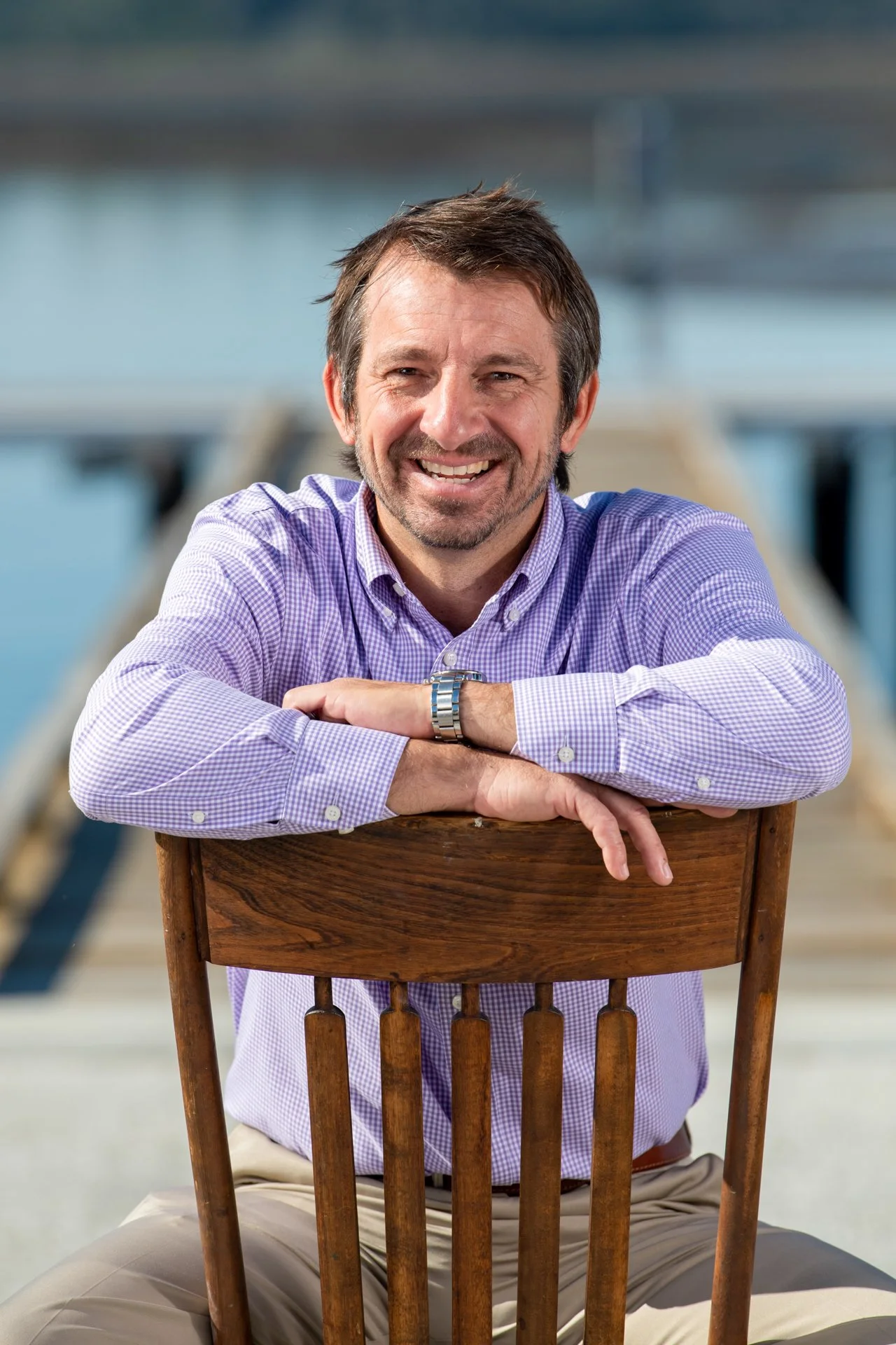 A middle-aged man with brown hair and a beard, smiling, sitting outdoors at a wooden chair with a dock in the background.