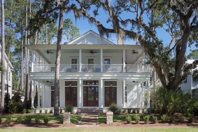 Two-story white house with a large front porch, surrounded by trees and small bushes, with a pathway leading up to the front steps.