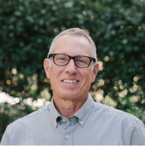 Eric Moser - Moser Kelly Design Group. Portrait of a man wearing glasses and a light blue button-up shirt outdoors with green foliage in the background.
