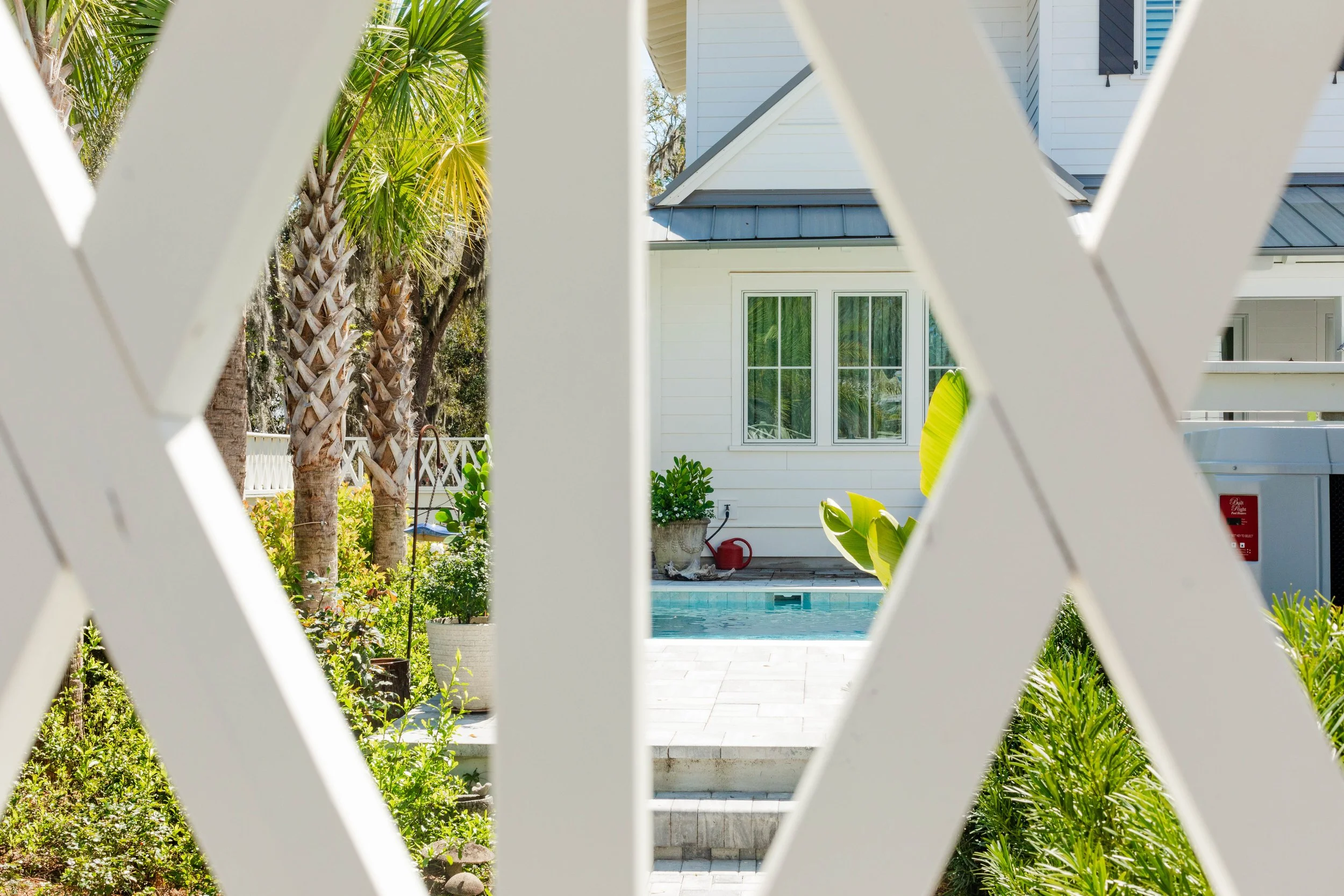 View of a backyard with a small swimming pool, surrounded by tropical plants and trees, seen through a white lattice fence. There is a house with white siding and windows in the background.