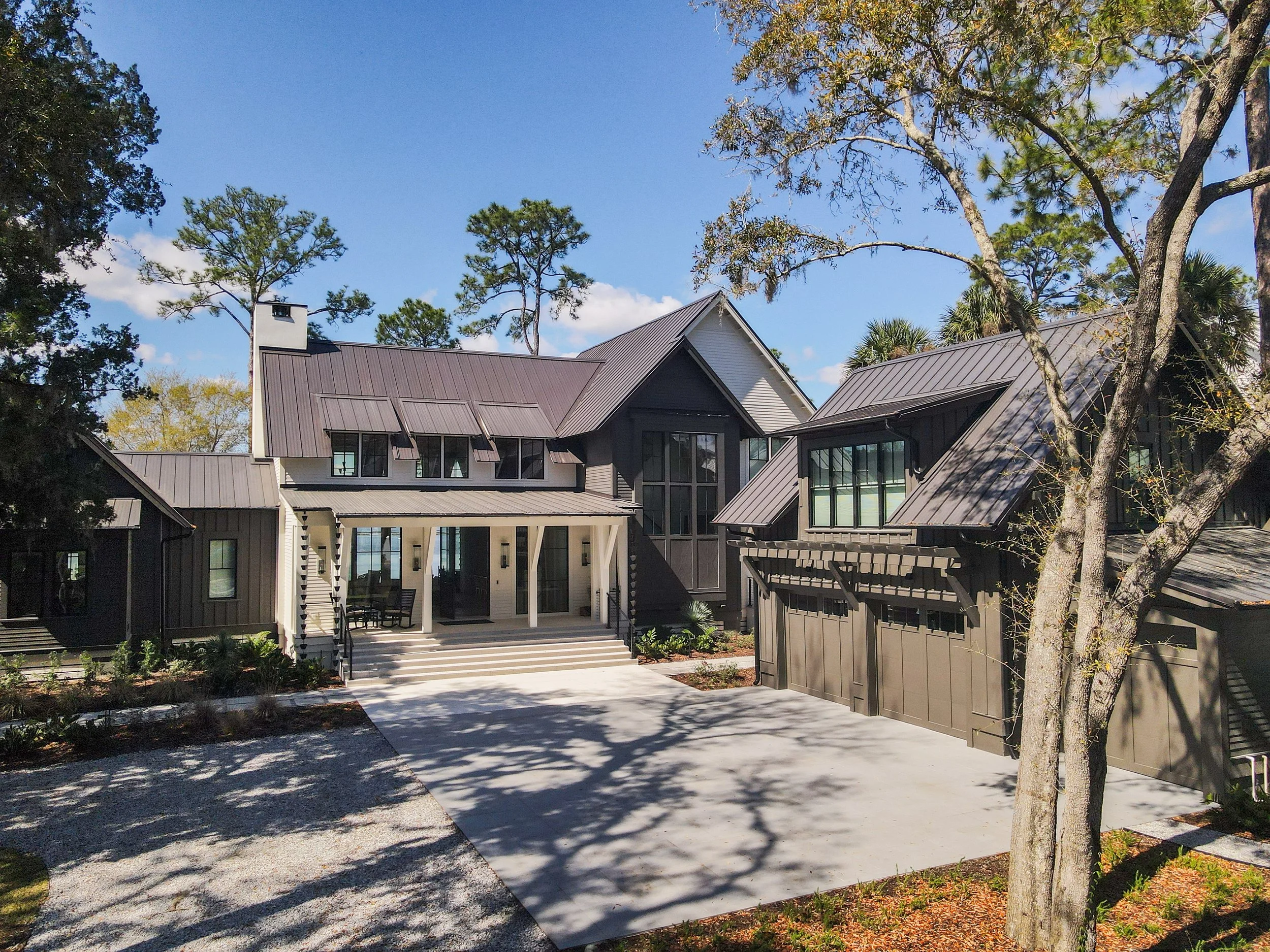 Moser Kelly Design Studio - Crane Island Intracoastal Home. A modern house with a metal roof, large windows, and a front porch with steps, surrounded by trees and landscaping in a rural area.