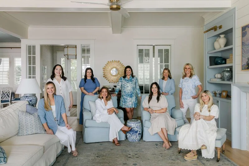 Lola Interiors Crane Island. Group of nine women gathered in a bright, stylish living room with white walls, blue and white decor, and a large mirror on the wall.
