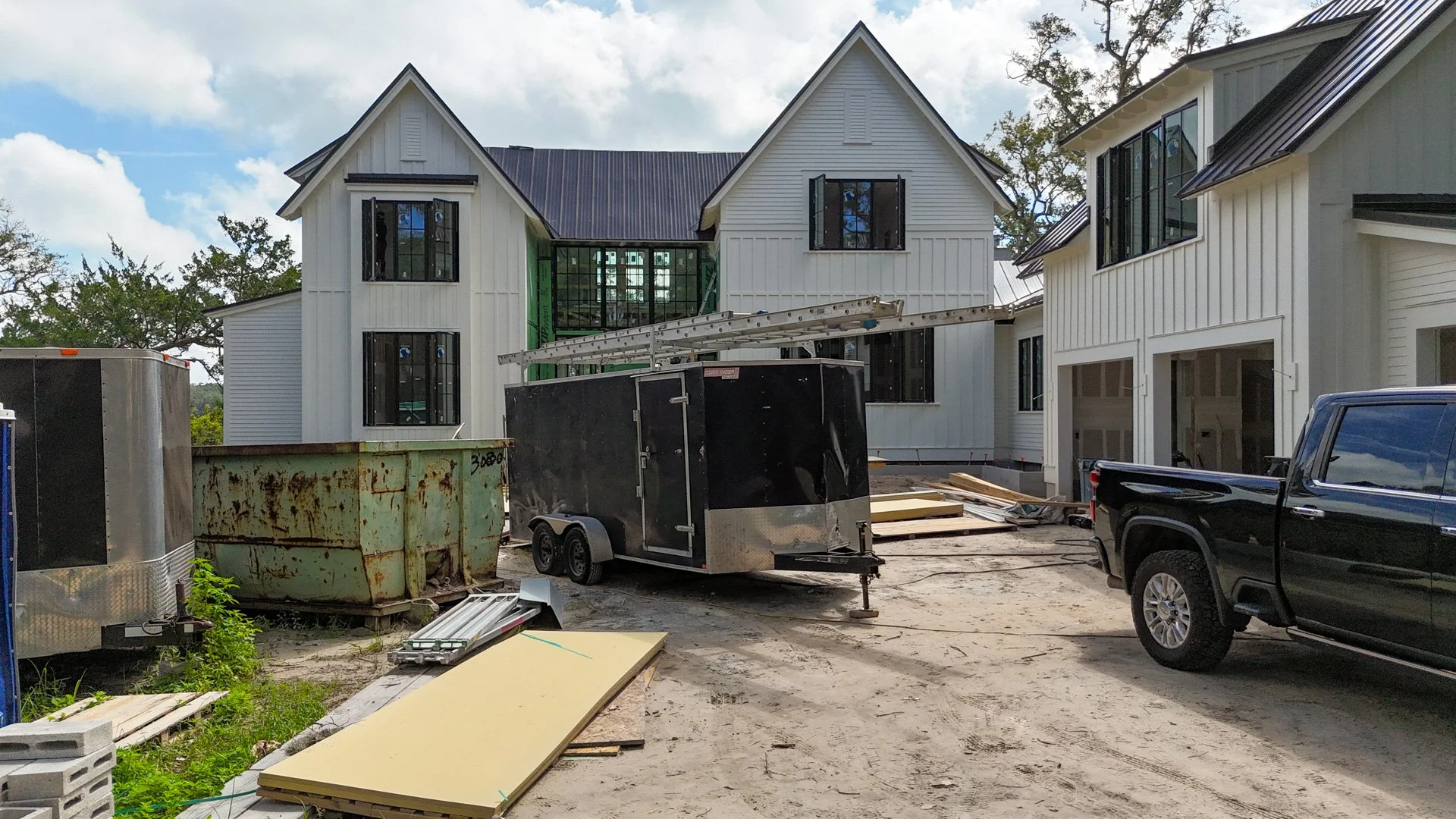 Construction site of a modern white house with black window frames, surrounded by construction equipment, trailers, and building materials.