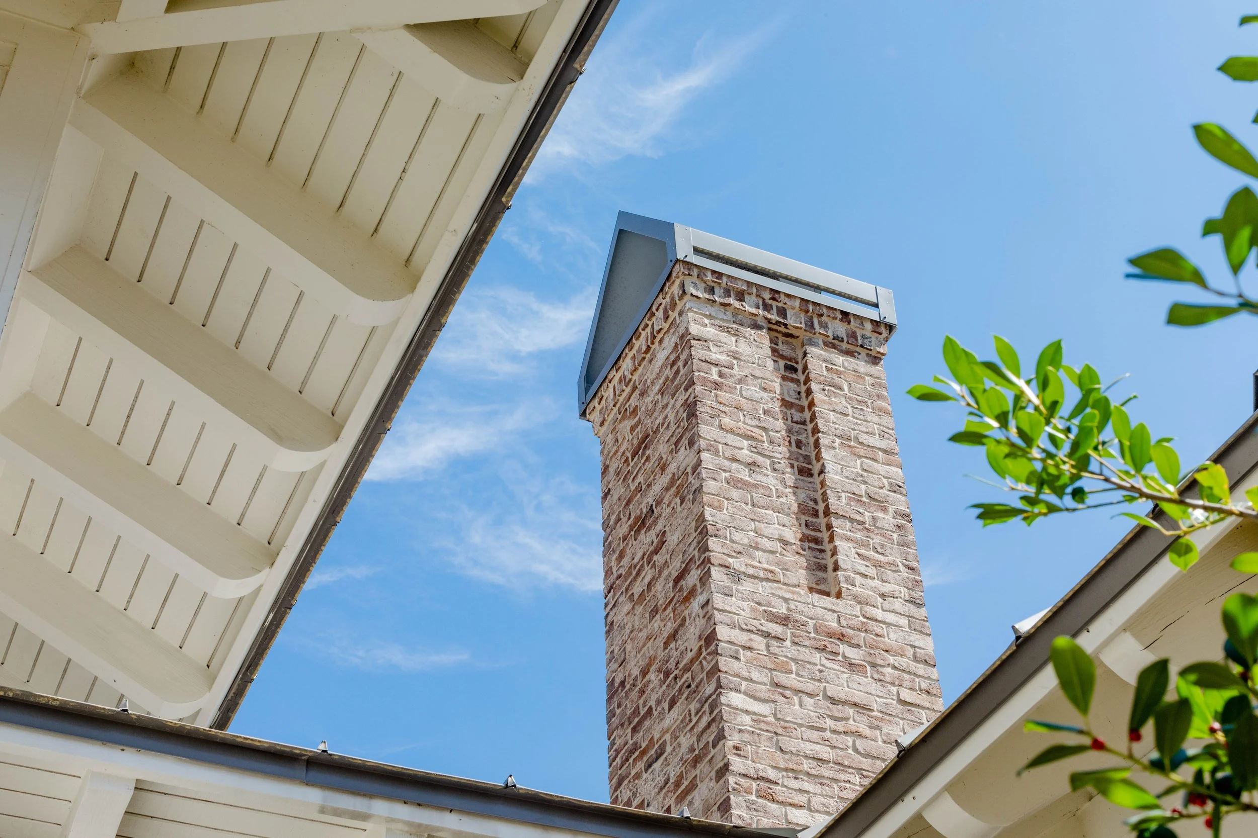 View of a brick chimney with a metal cap, seen from below between the eaves of a house, under a clear blue sky with some wispy clouds and green leaves in the foreground.