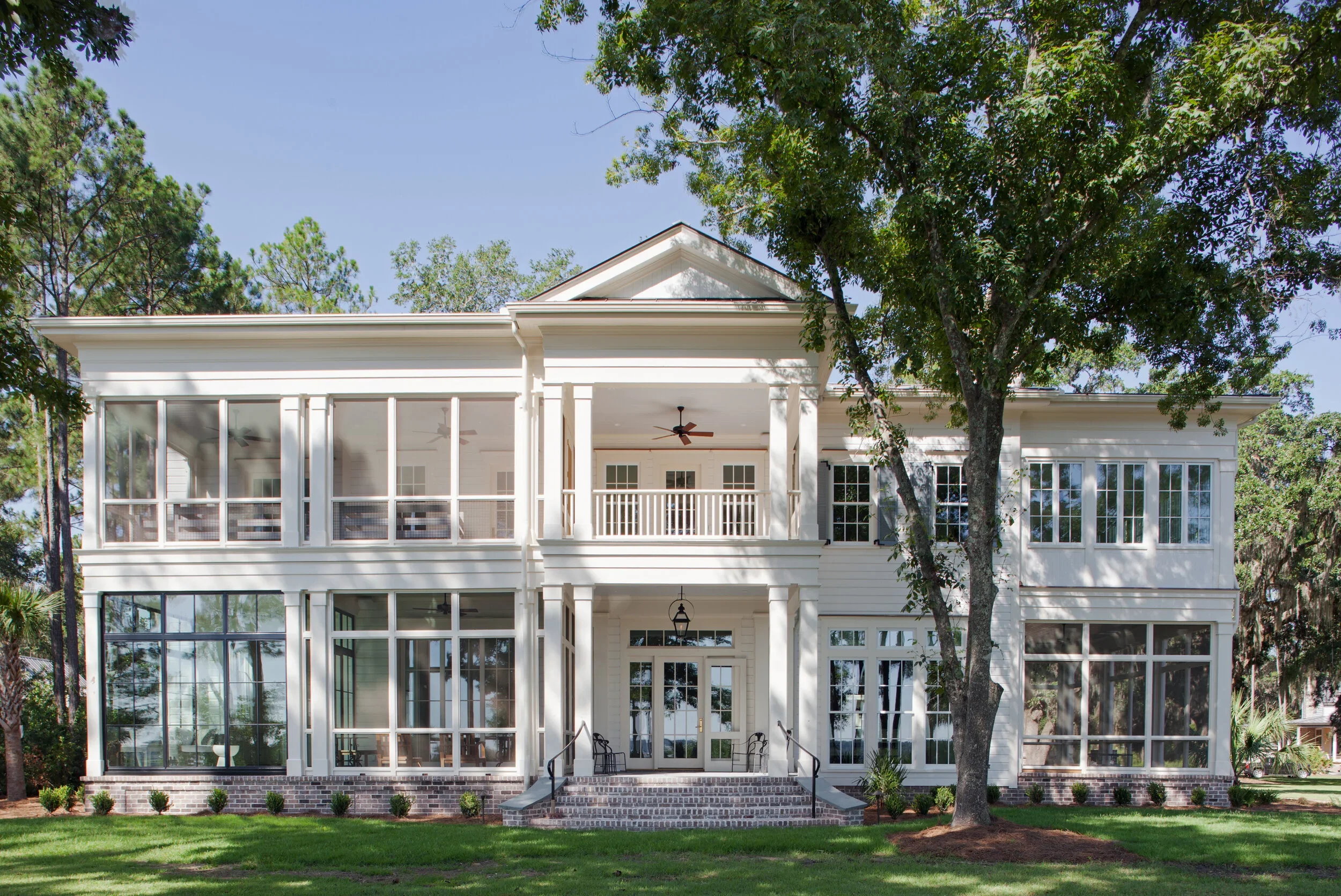 Hansen Architecture - Crane Island, FL. A large two-story white house with a screened-in porch on the upper level, multiple windows, and a brick staircase leading to the front entrance, surrounded by a green lawn and trees.