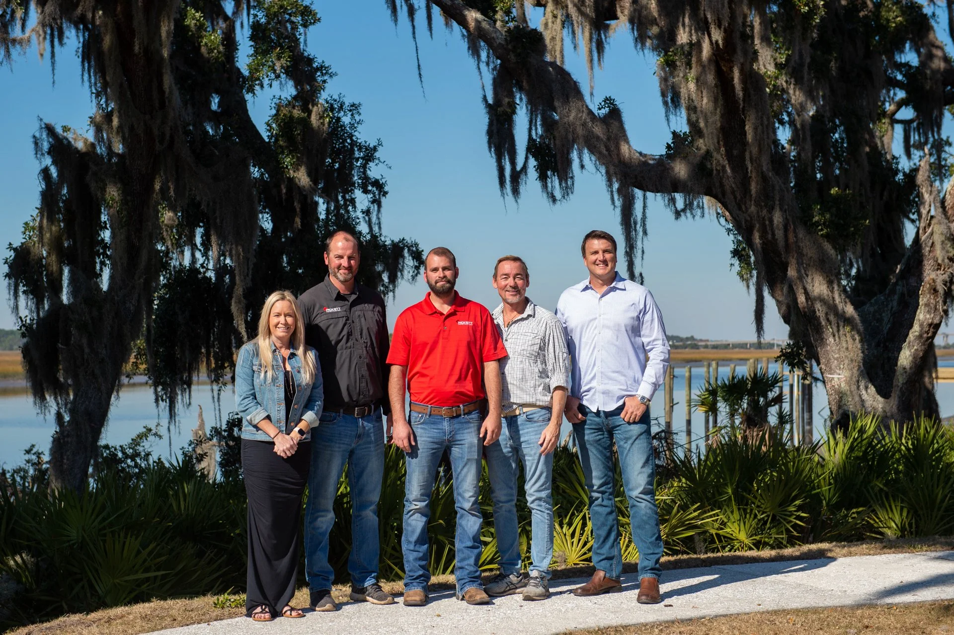 Group of five people standing outdoors near water and trees, smiling for the camera.
