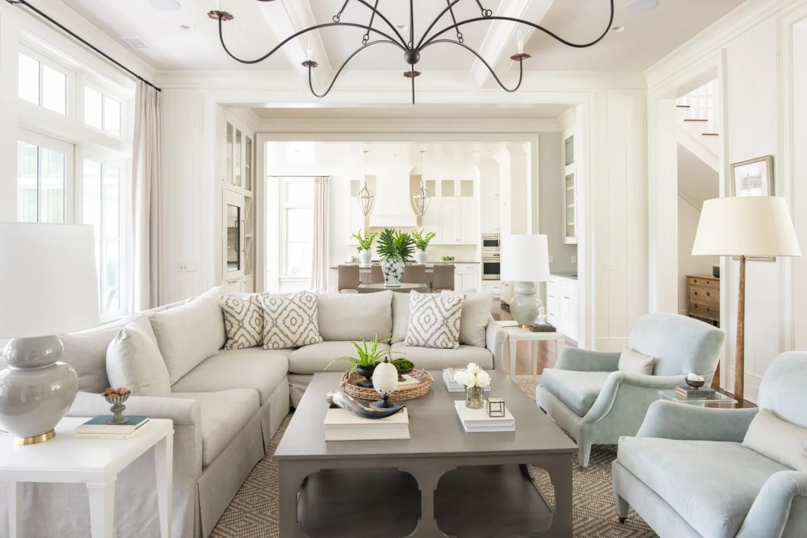 Bright, airy living room with white walls, large windows, and neutral furniture including a beige sectional sofa and blue armchairs. A coffee table holds decorative items and plants, with a modern chandelier hanging from the ceiling. The open floor p