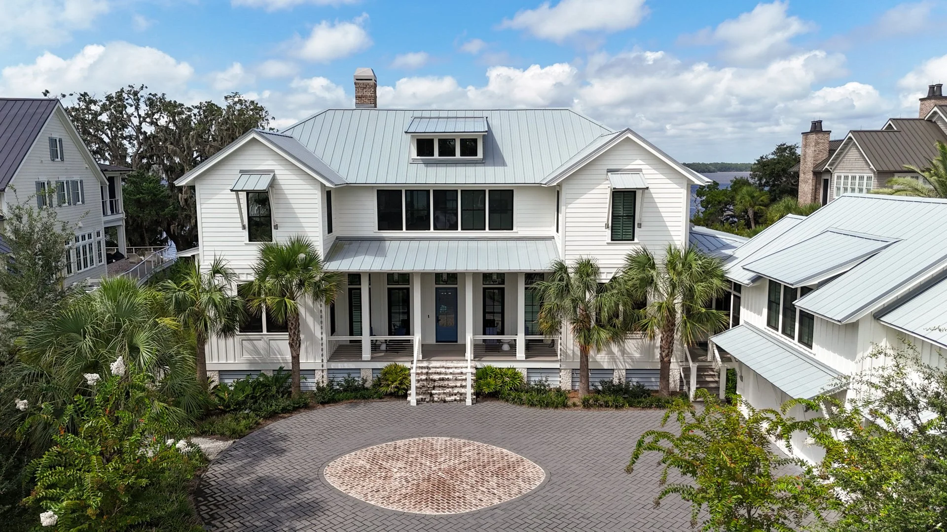 A white, three-story house with a metal roof, surrounded by palm trees and a paved circular driveway in front. Other houses with similar styles are visible on either side, and there is a body of water in the background under a partly cloudy sky.
