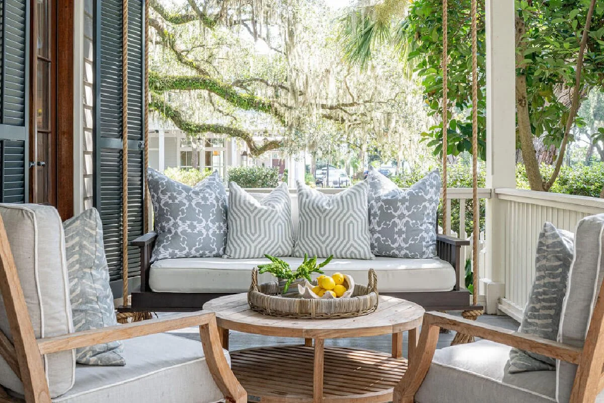 Outdoor porch with white cushioned furniture, grey and white patterned pillows, wooden table with a tray of lemons and greenery, surrounded by green trees and plants, on a sunny day.
