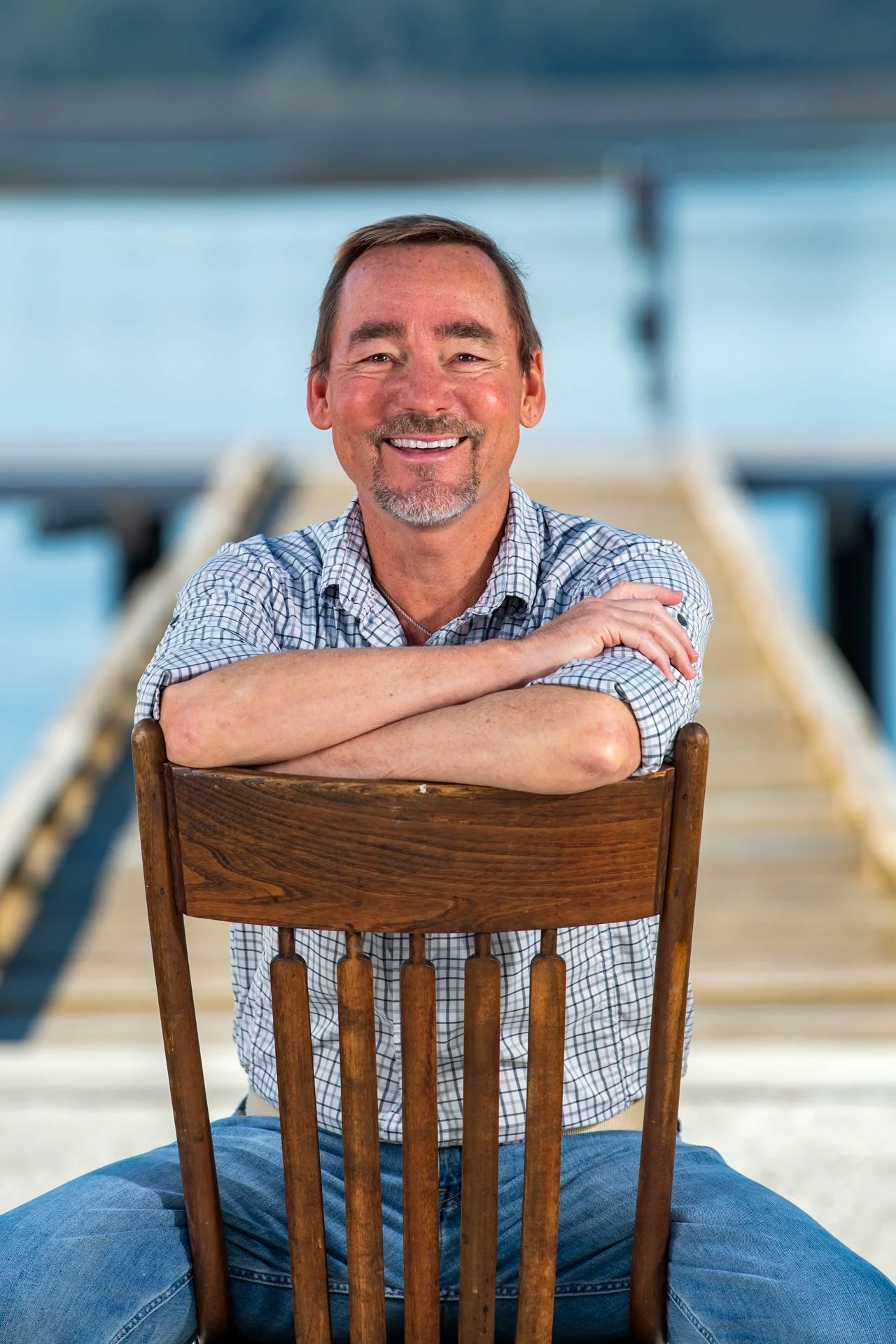 A smiling middle-aged man with short dark hair, a beard, and wearing a plaid shirt, sitting behind a wooden chair on a dock near the water.