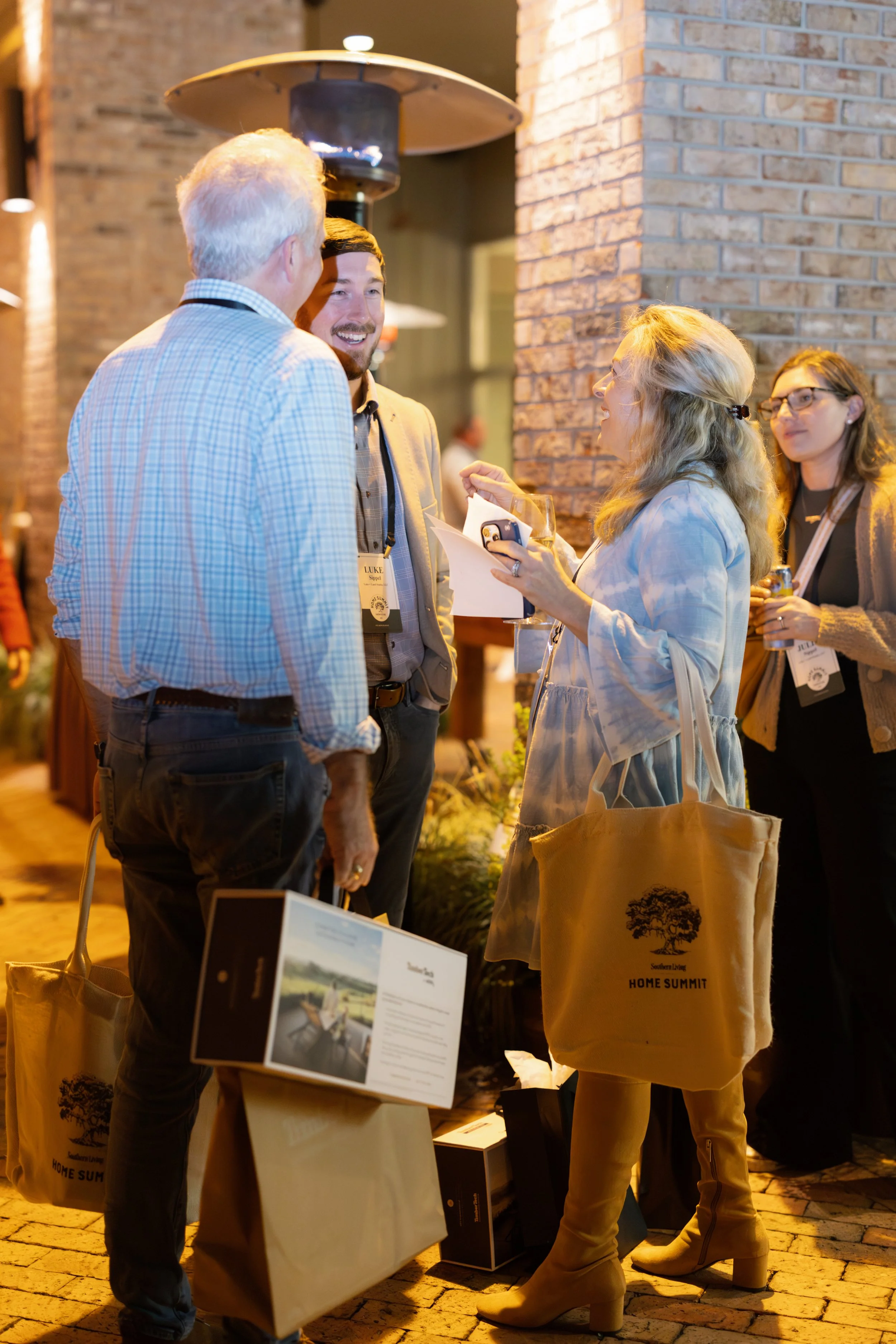 John Hillman & Dawn Hillman Cabana Lane. Four people engaged in conversation at a networking event, holding tote bags and brochures, with a brick wall and outdoor heater in the background.