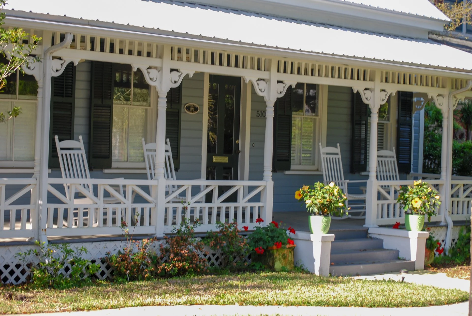 A house with a porch featuring white trim, two rocking chairs, and potted yellow and white flowers on the steps, with a small garden in front. Downtown Fernandina Beach, FL - Examples of Lowcountry Architecture.