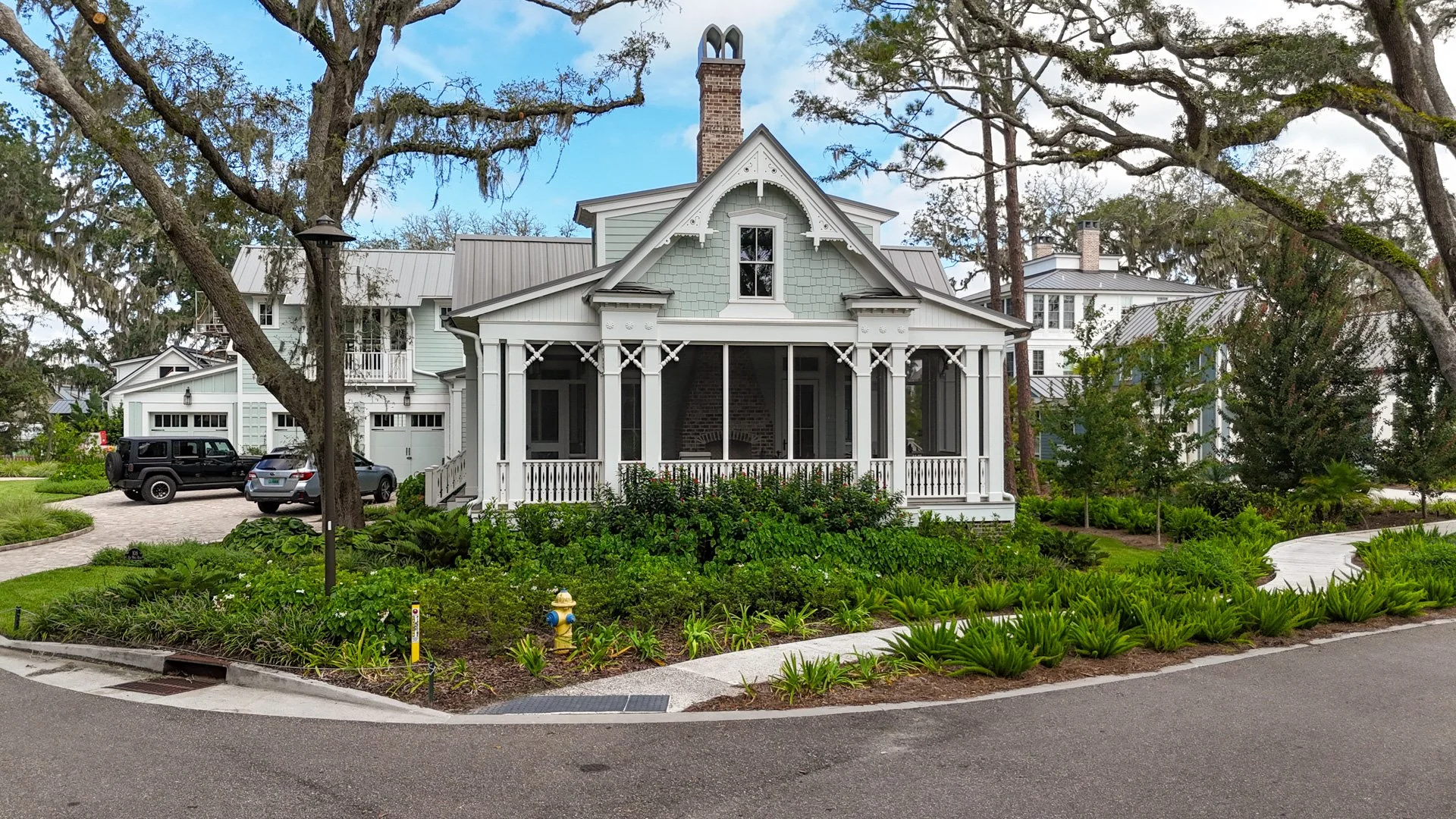 Historical Concepts - Crane Island Homestead Collection. A charming white house with a screened porch, surrounded by lush landscaping and trees, with a driveway and parked cars.