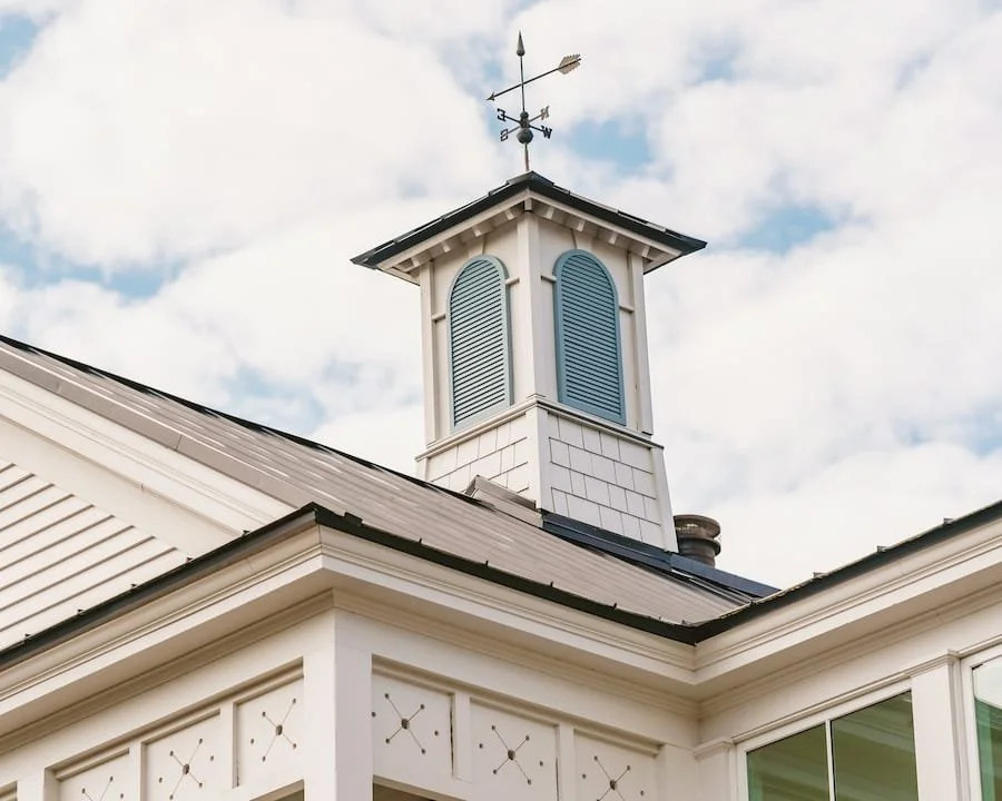 Close-up of a white house's cupola with blue shutters, a weather vane on top, and part of the roof and window visible under a partly cloudy sky.