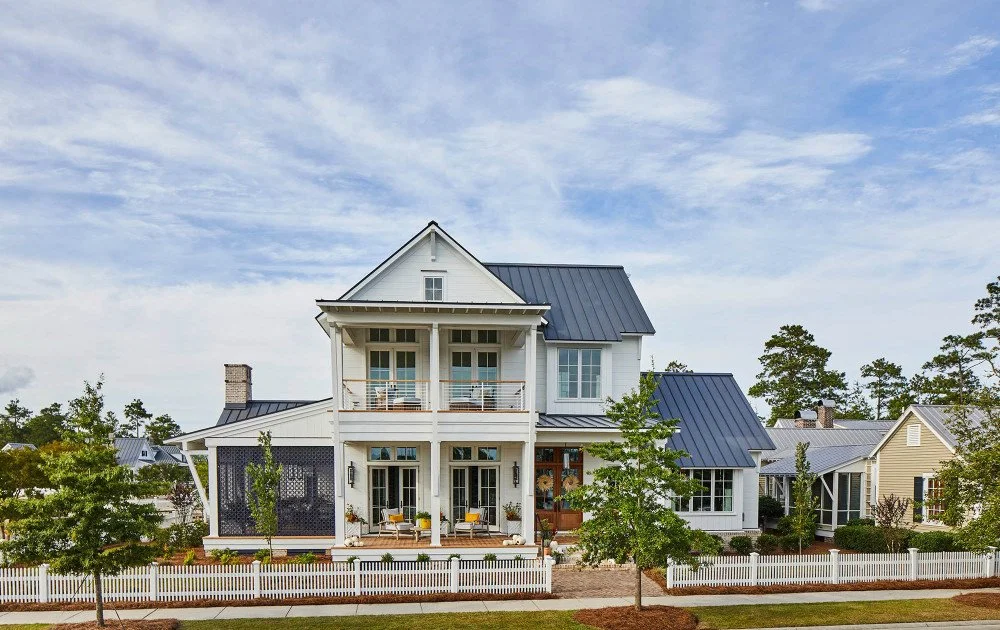 A modern two-story white house with a blue metal roof, front porch, and balcony, surrounded by a white picket fence and greenery.