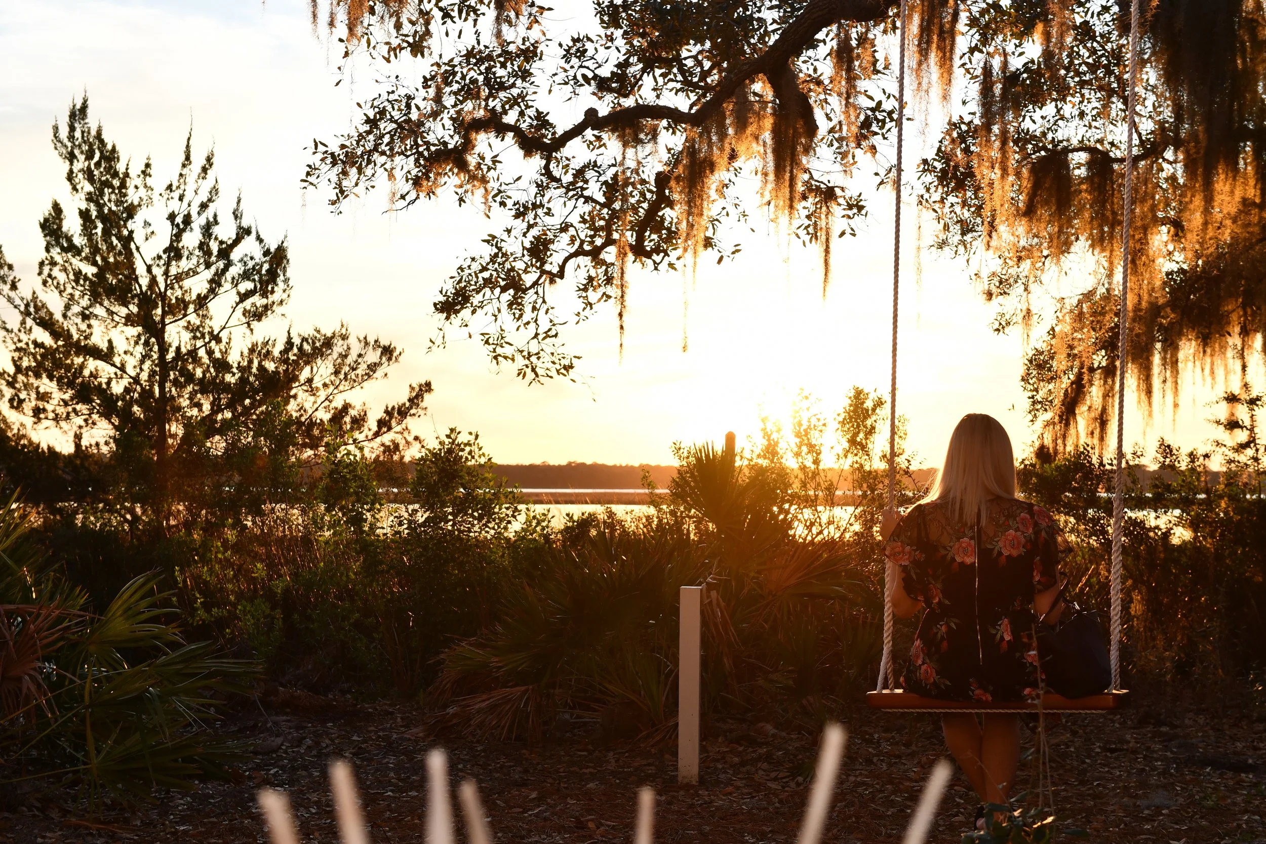 2019 Southern Living Idea House. A woman with blonde hair sitting on a swing at sunset, surrounded by trees and bushy plants near a body of water.