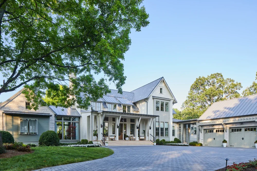 Lake & Land Studio - Southern Living Idea House. Large white house with a front porch, surrounded by a green lawn and trees, with a paved driveway leading to a three-car garage.