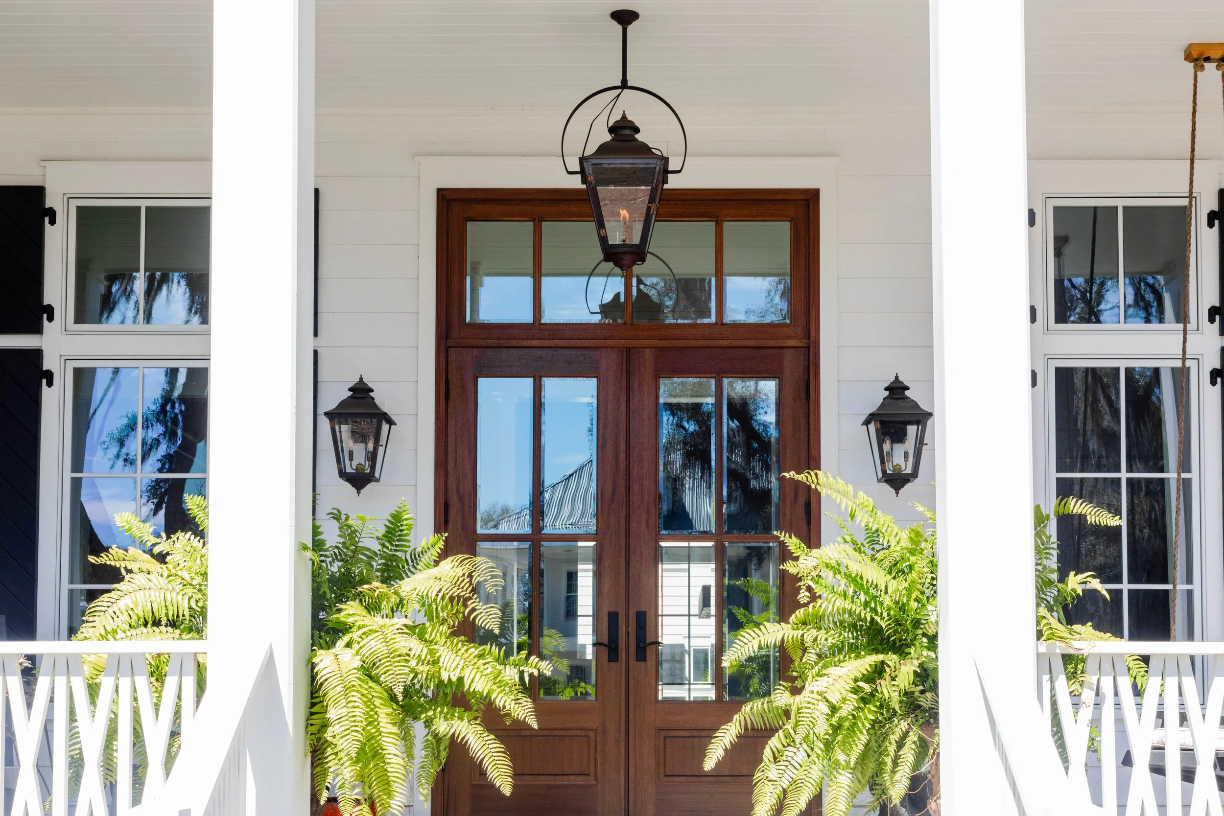 Front porch of a house with a large wooden door, two vintage black lantern wall lights, hanging and potted ferns, and two glass-paneled windows.