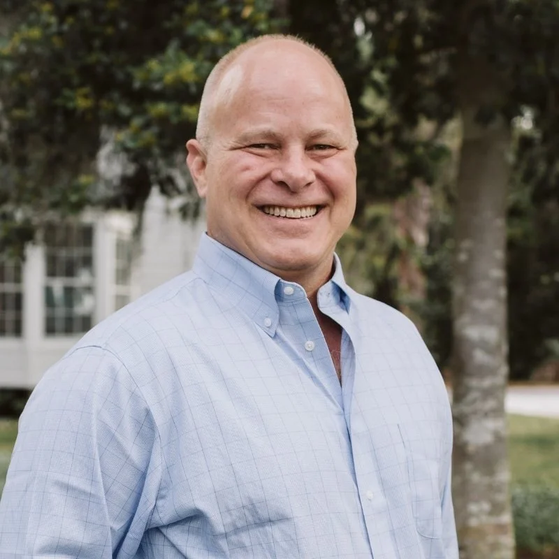 Perce Scott Architects - Pearce Scott - Crane Island. A smiling bald man in a blue button-up shirt standing outside in front of trees and a house.